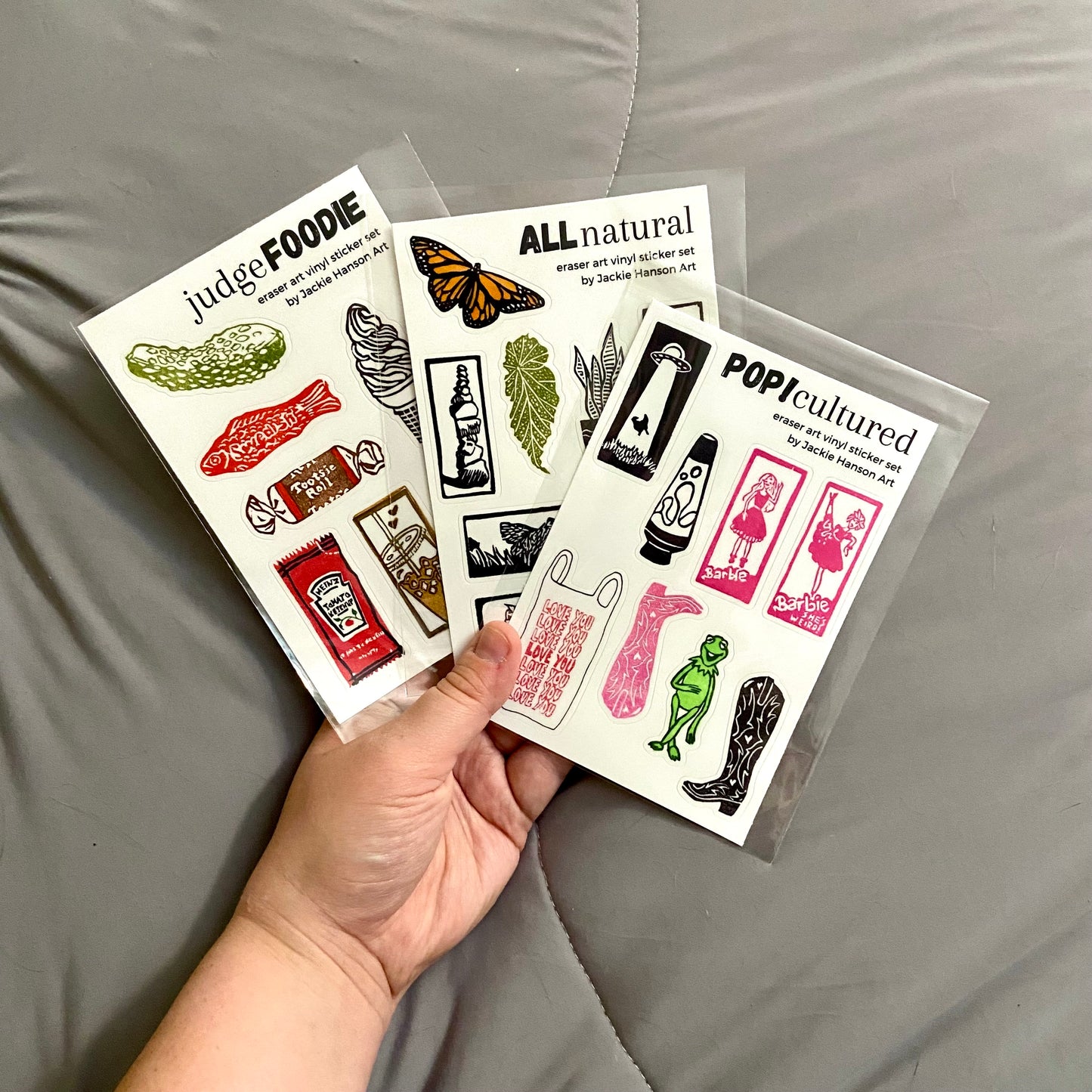 The artist's outstretched hand fans out all three varieties of her eraser print sticker sets: judgeFOODIE, ALLnatural, and POP!cultured. Between the three sets, there are 23 quality vinyl stickers of her small relief prints.