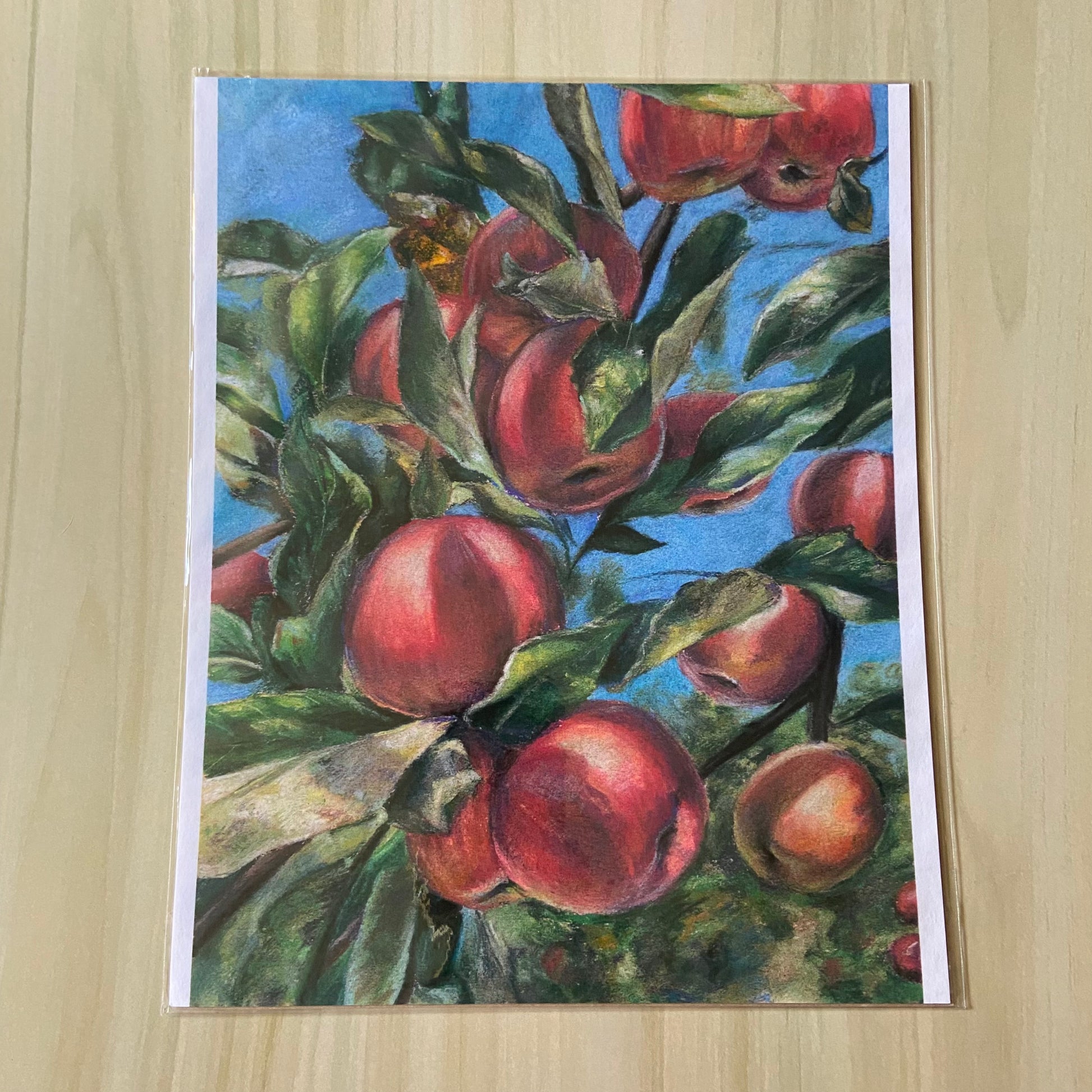 An art print of red apples growing on a tree. The print focuses on three main fruits with several others visible in the midground. Leaves from the apple tree are woven throughout and a blue sky with a few trees is behind.
