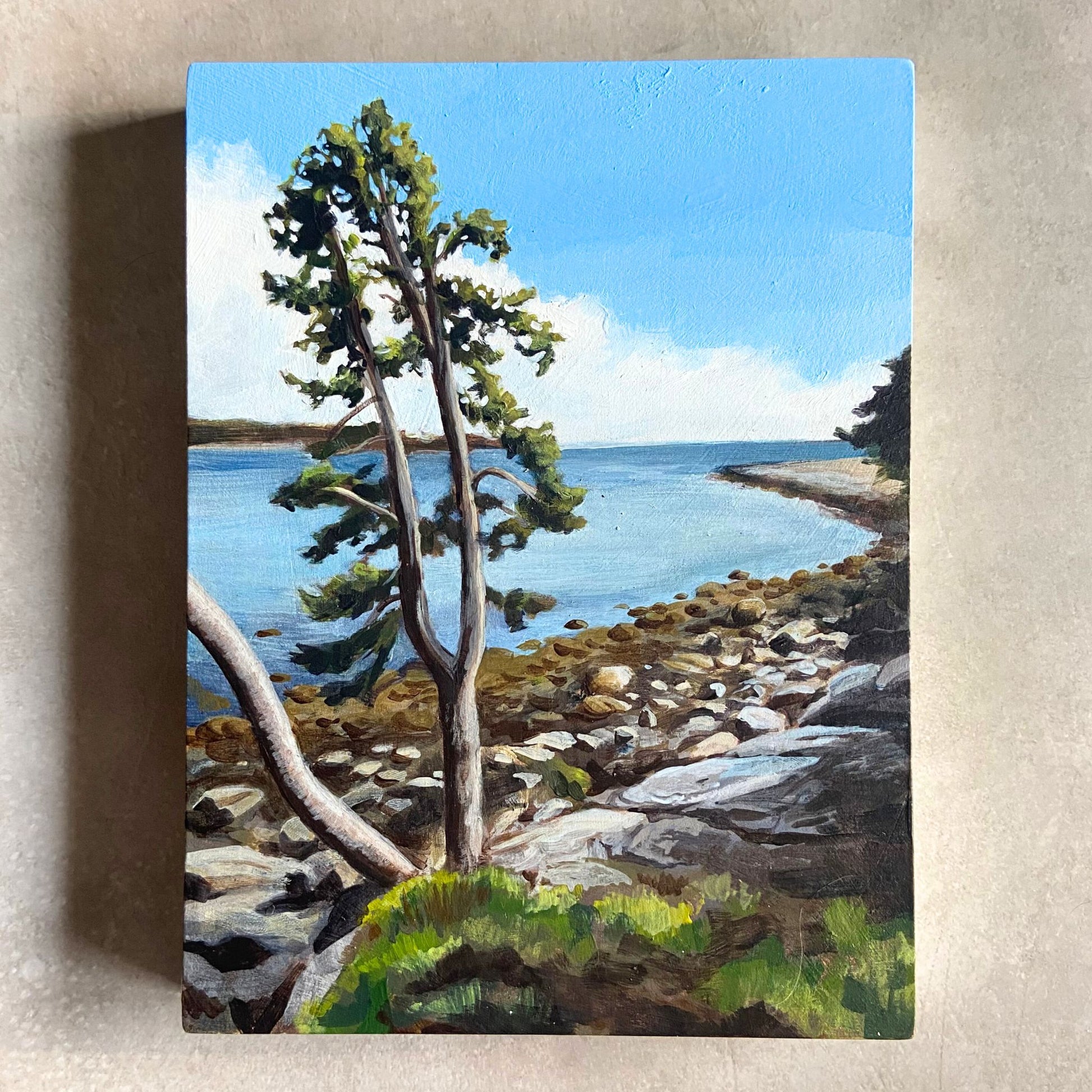 An acrylic coastal landscape painting on wood panel sitting on a textured background. the painting, portrait-oriented, depicts a bay on the ocean on a blue-skied day. The beach is rocky and the focal point is a tree growing at the edge of the land.