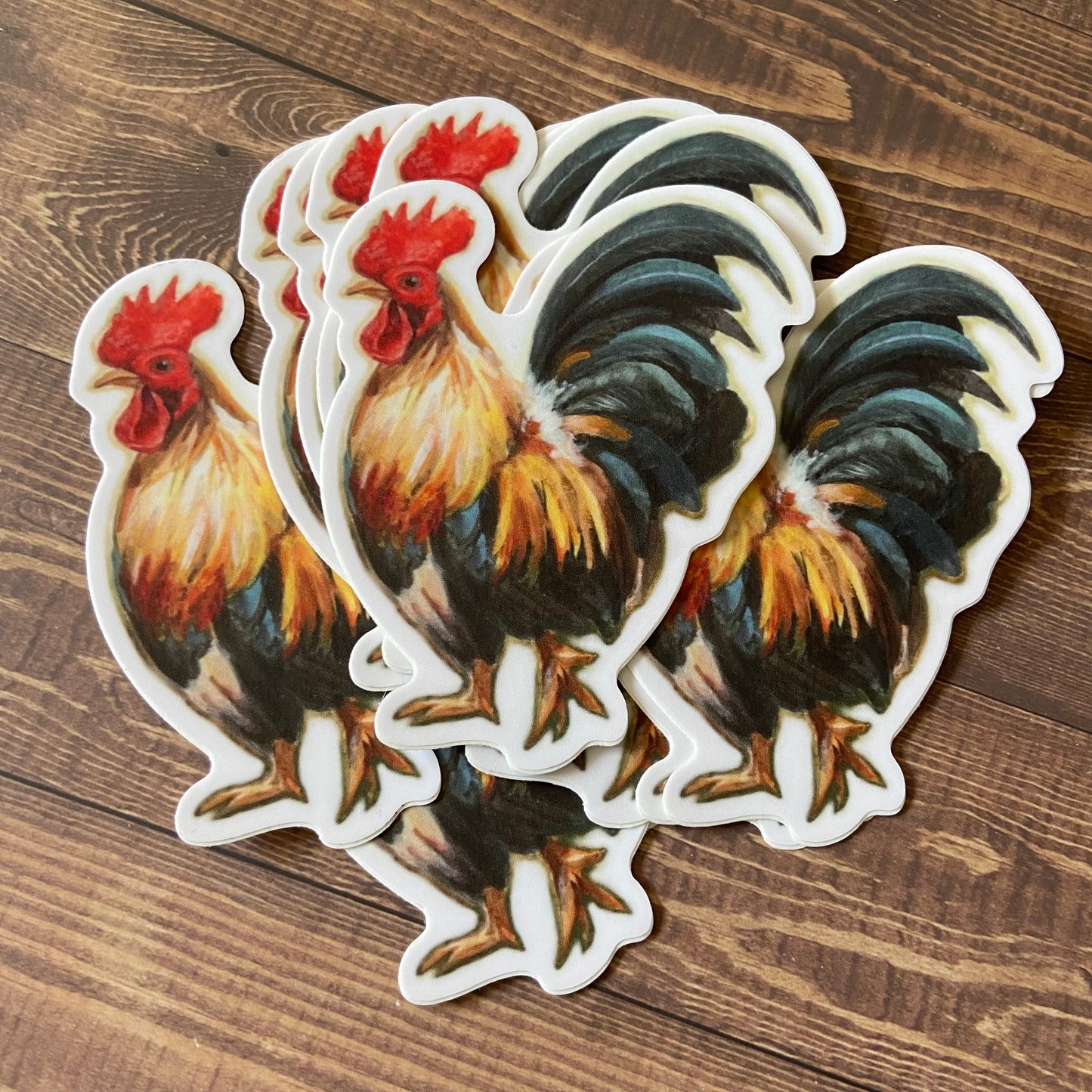 A small pile of rooster stickers on a wood surface. The roosters are seen from the side, with bright multicolored feathers and a calm expression. He's got one foot raised in mid-stride. Each decal has a thin white border.
