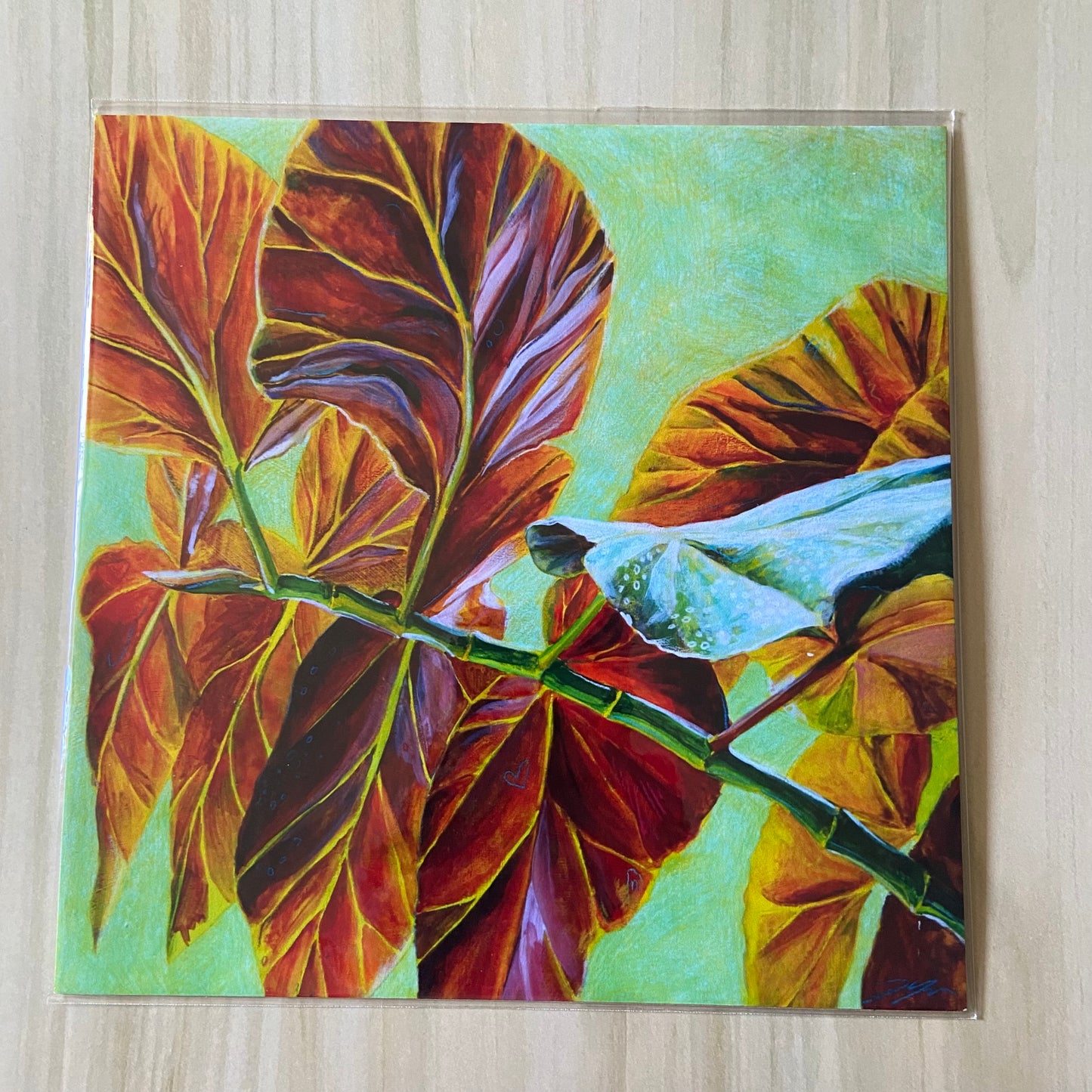 A square art print of red leaves over a pale green background. They stem from a branch starts at bottom right and goes up almost to center left. One leaf shows part of its other side, green with white spots. The print is on a wood surface.