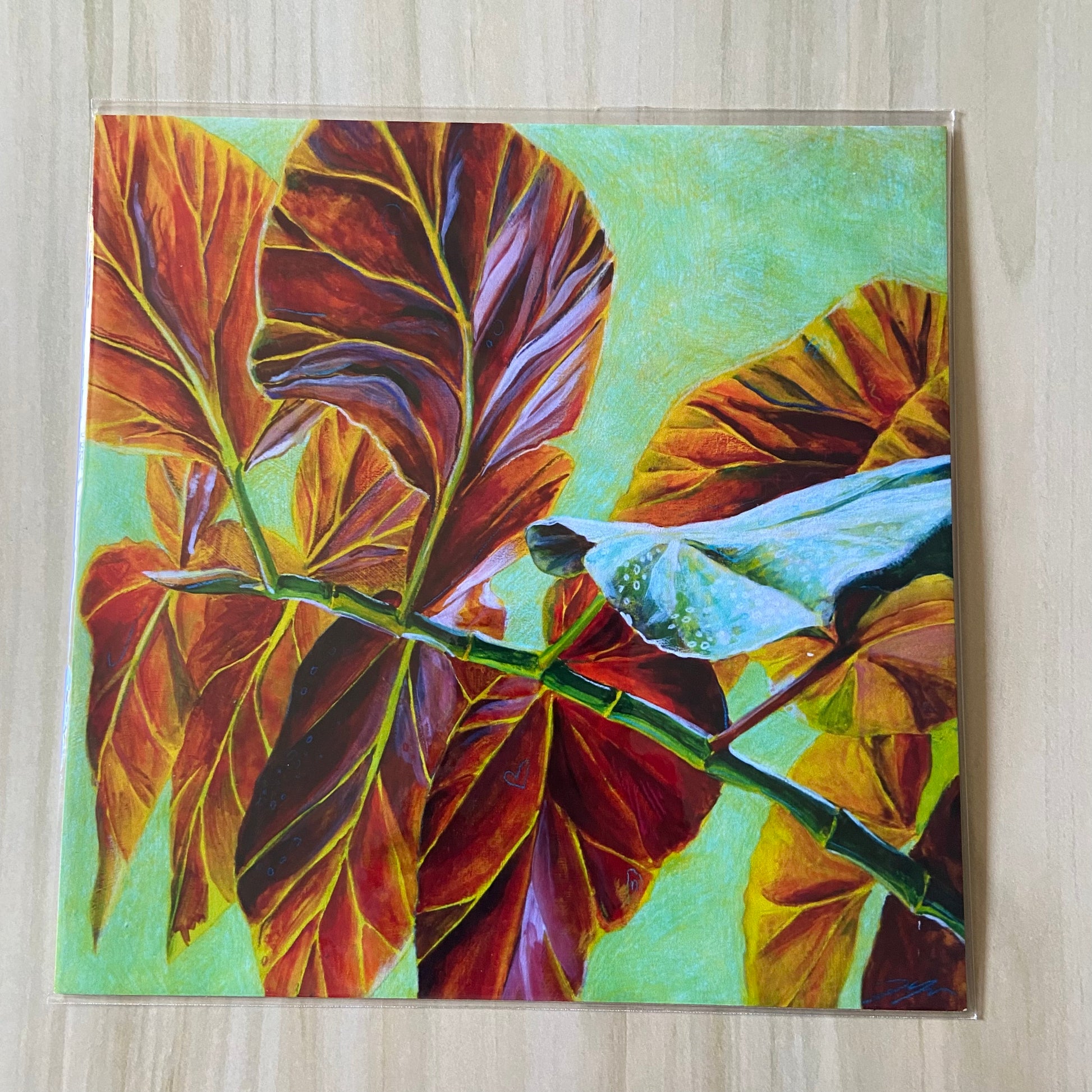 A square art print of red leaves over a pale green background. They stem from a branch starts at bottom right and goes up almost to center left. One leaf shows part of its other side, green with white spots. The print is on a wood surface.