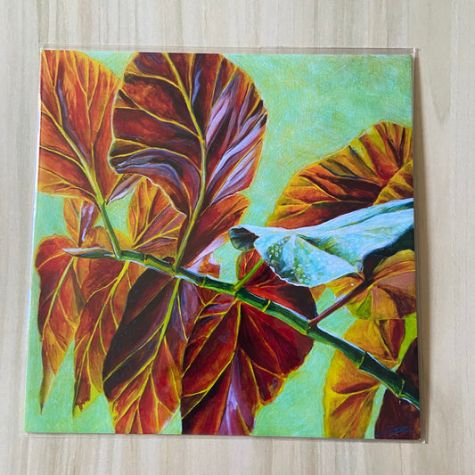 A square art print of red leaves over a pale green background. They stem from a branch starts at bottom right and goes up almost to center left. One leaf shows part of its other side, green with white spots. The print is on a wood surface.