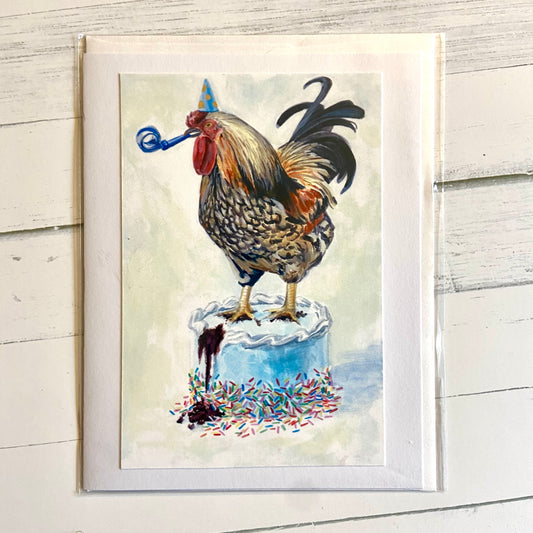 A folded white greeting card with an image of my gouache painting, Birthday Rooster. A rooster stands on top of a blue birthday cake, with a blue noisemaker in his beak and wearing a blue party hat.