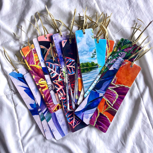 A variety of bookmarks fanned out on white fabric. They are in many colors, all with a gold ribbon tied at the top.