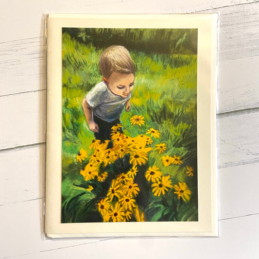 A folded cream-colored greeting card with an image on the front of a painted little boy looking down at some Black-eyed-Susan flowers in a lush green area. the painting was made with pastels, resulting in a chalky texture.
