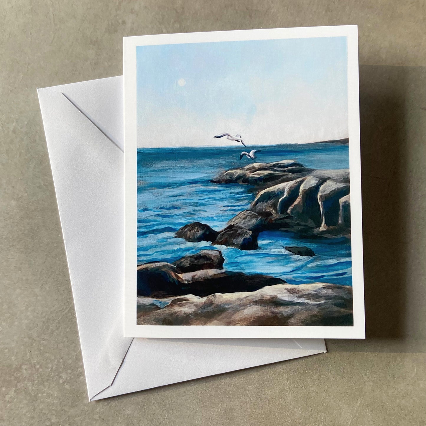 A folded white greeting card and envelope on a grey surface. The card shows a painting of rocks at sea with two gulls flying near the horizon line.