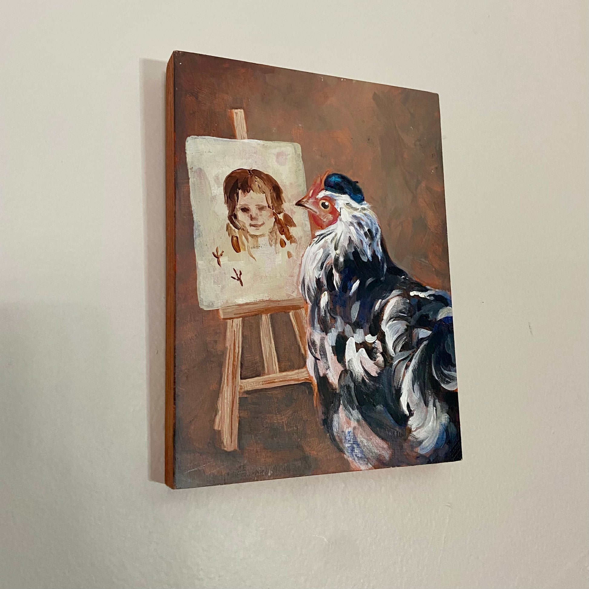 A rectangular painting of a black and white chicken, wearing a black beret, standing in front of an easel, presumably painting. The easel has a crude portrait of a human woman and two small chicken footprints (a signature). The background is abstract, textured brown. The painting hangs on a light gray wall.