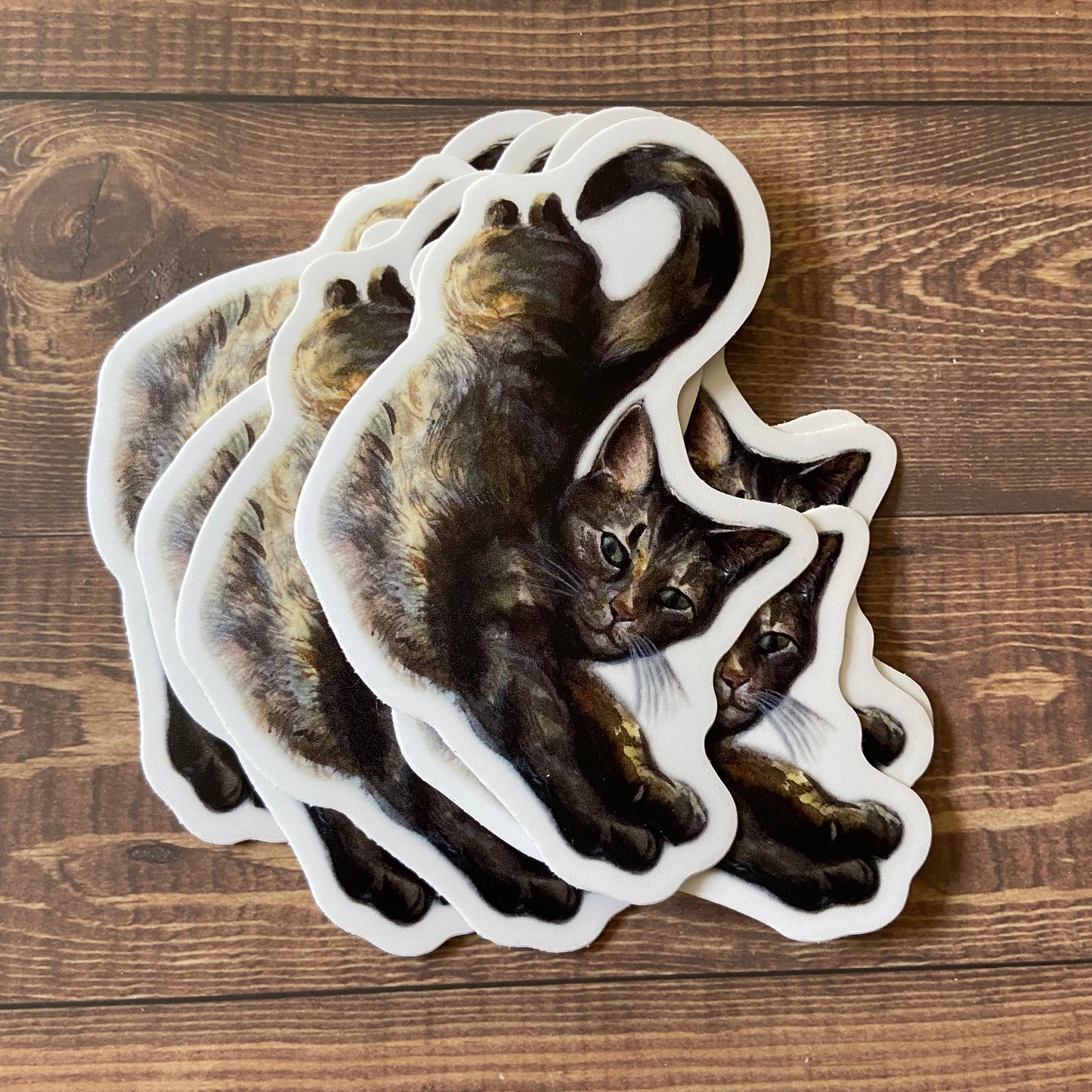A small pile of stickers, all depicting a watercolor cat painting. The feline is laid out, stretching long toward the viewer. She is very fluffy and relaxed, with a tail curling in the air. Stickers have a thin white border and are on a wood surface.