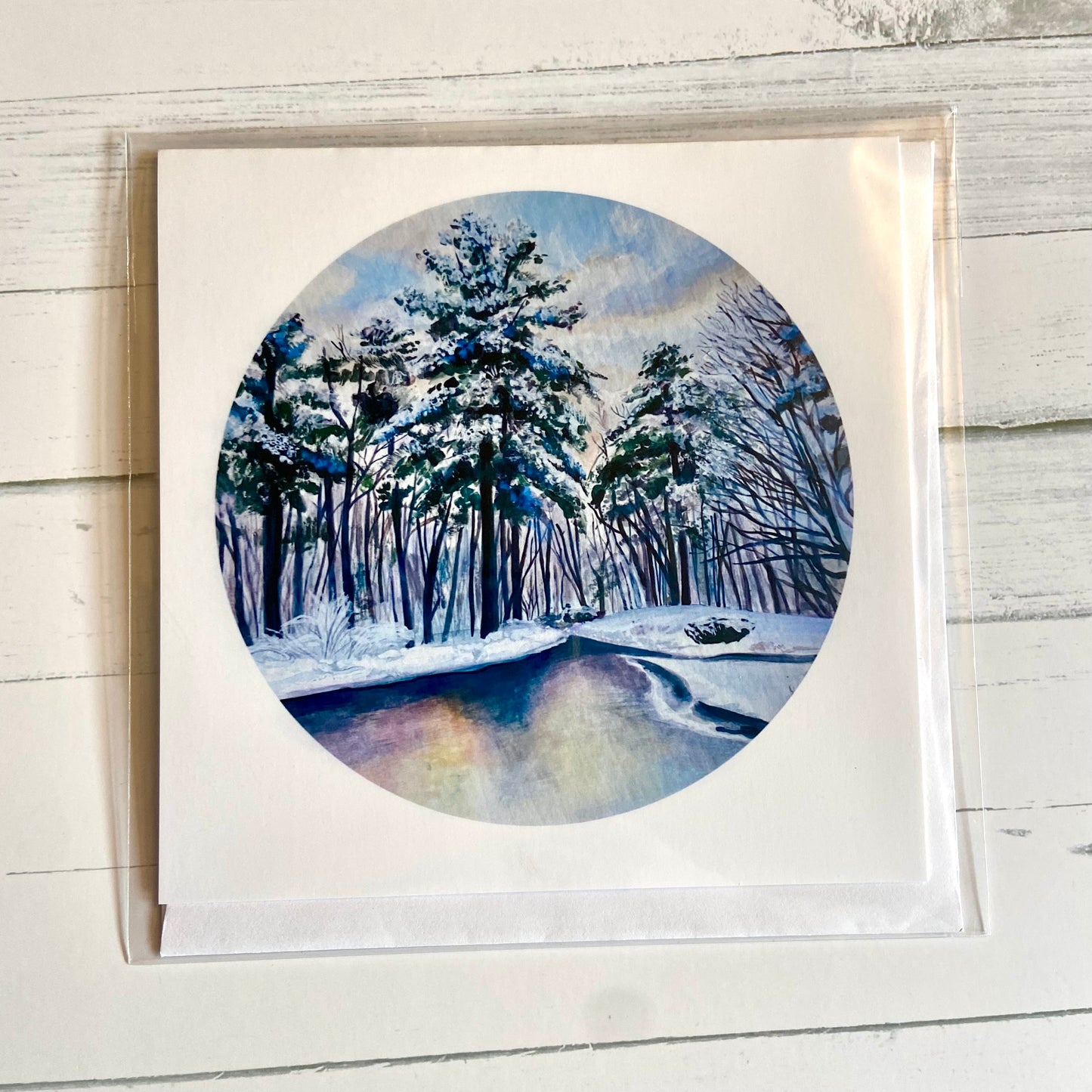 A folded white, square greeting card featuring my round painting, December 26, on the front. The painting is a predominantly blue snowy landscape, with snowy pines lining a still river.