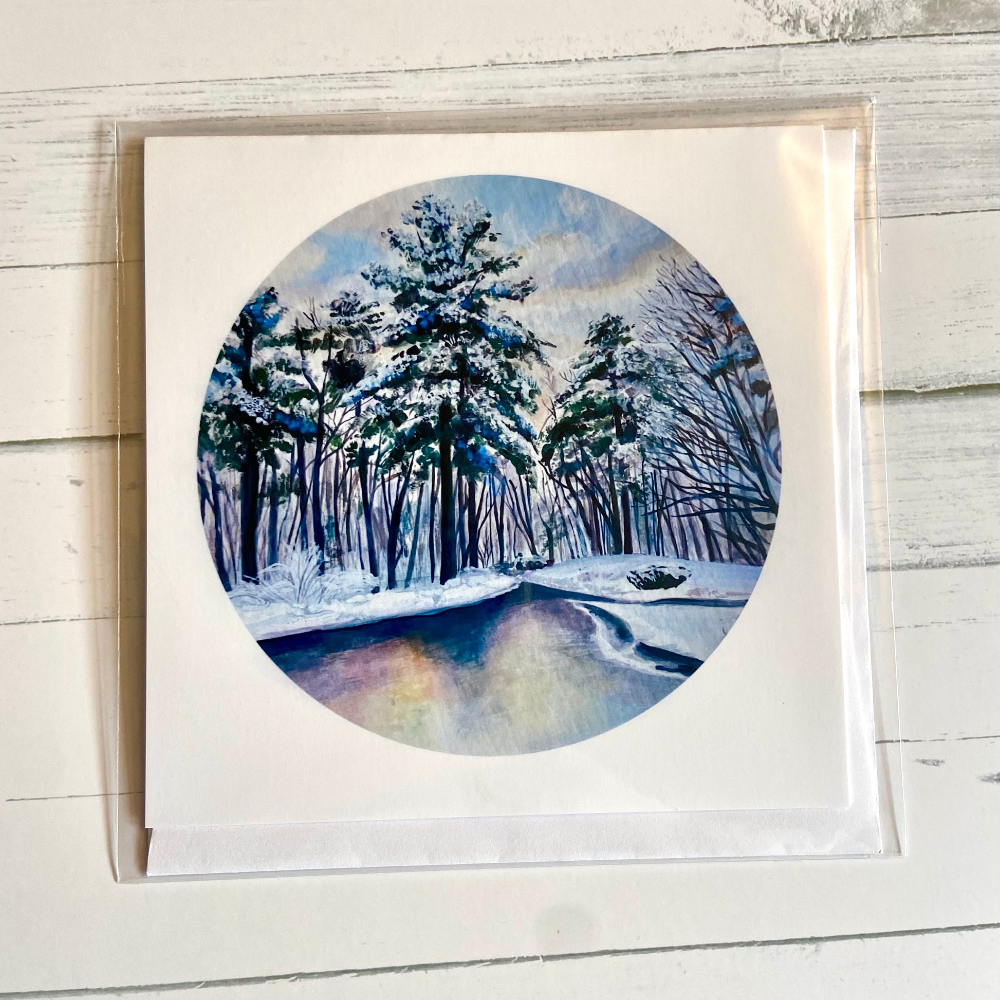 A folded white, square greeting card featuring my round painting, December 26, on the front. The painting is a predominantly blue snowy landscape, with snowy pines lining a still river.