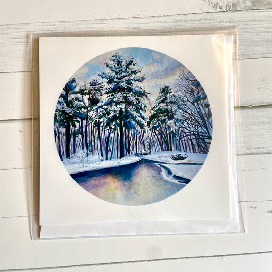A folded white, square greeting card featuring my round painting, December 26, on the front. The painting is a predominantly blue snowy landscape, with snowy pines lining a still river.
