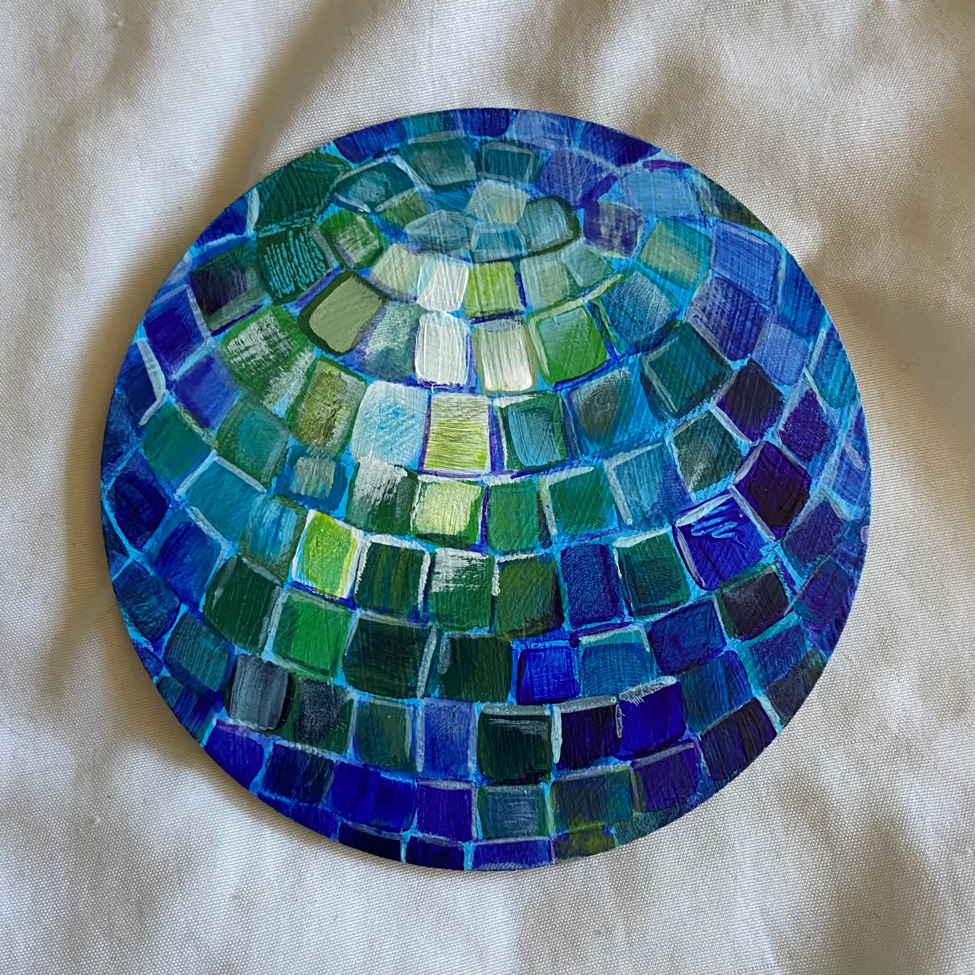 A small round painting of a disco ball, with blue mirrors in shadow and green/white highlights.