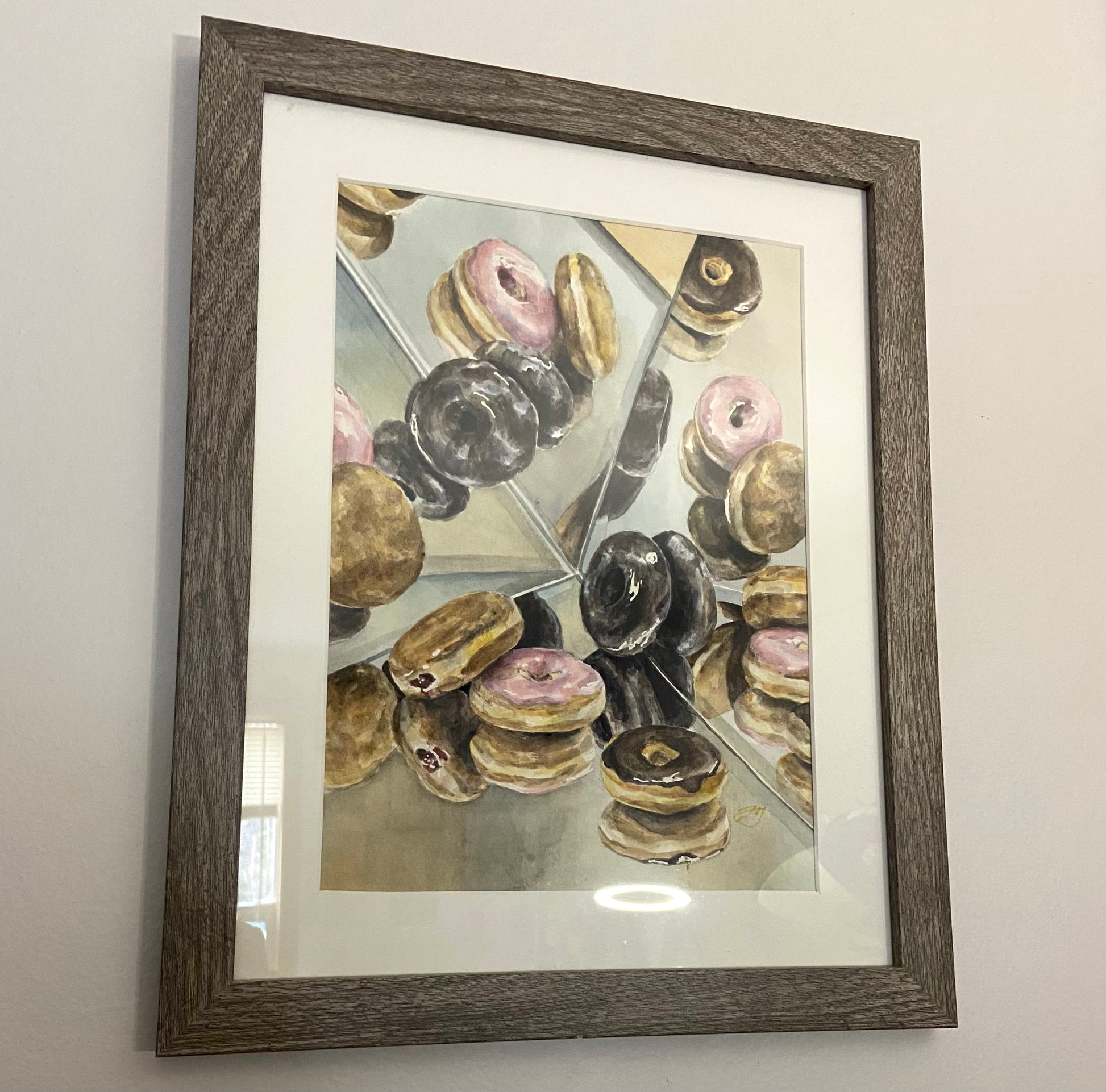 A watercolor still life painting of several varieties of donuts surrounded by mirrors, like a kaleidoscope. The painting is in a cool wood frame with a white mat.