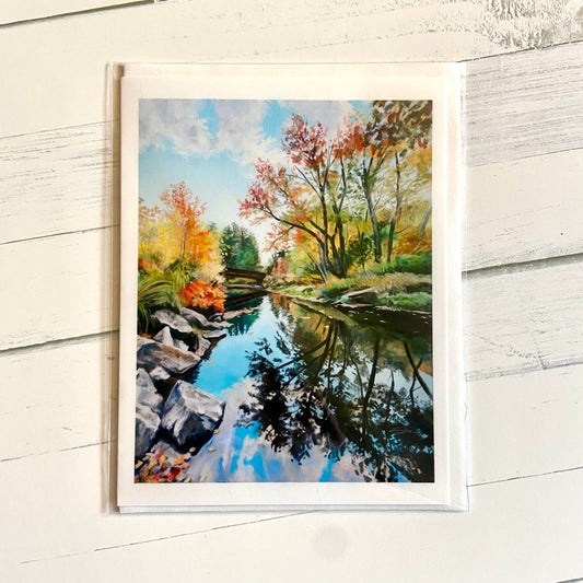 A folded white greeting card with an image of my soft pastel landscape painting, Elderglass. Shown is the mirrorlike Blackwatwer River from rural New Hampshire, with autumn colors in the trees surrounding. A bridge crosses the river in the distance, and several delicate trees dance over the water with a deep reflection underneath.