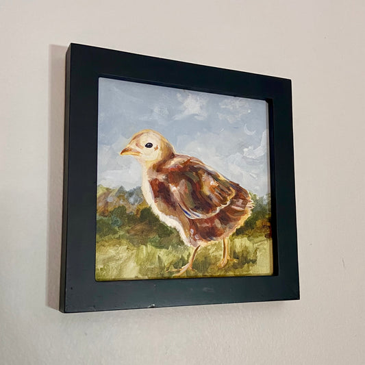 A small, square painting in a black frame on a gray wall. The painting depicts a yellow and brown baby chick walking to the left across a grass field.