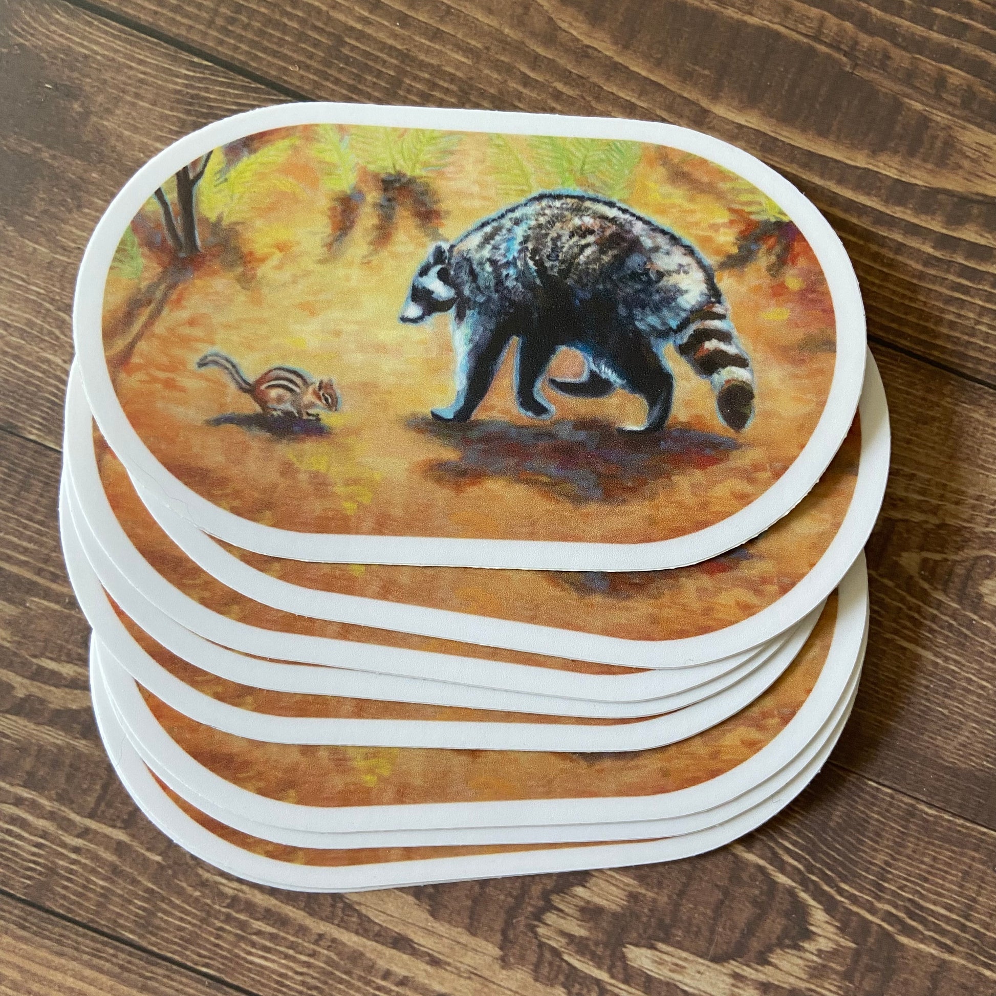 A small pile of orange, oval-shaped stickers on a wooden surface. The decals depict a gouache painting of a raccoon and a chipmunk making friends on the forest floor. Each sticker has a thin white border.
