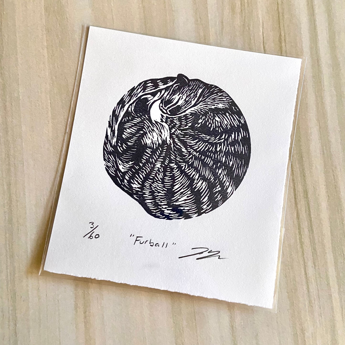 A circular black-and-white print of a striped cat curled into a circle for a nap. The print is made by hand using a carved eraser and is numbered, titled and signed.