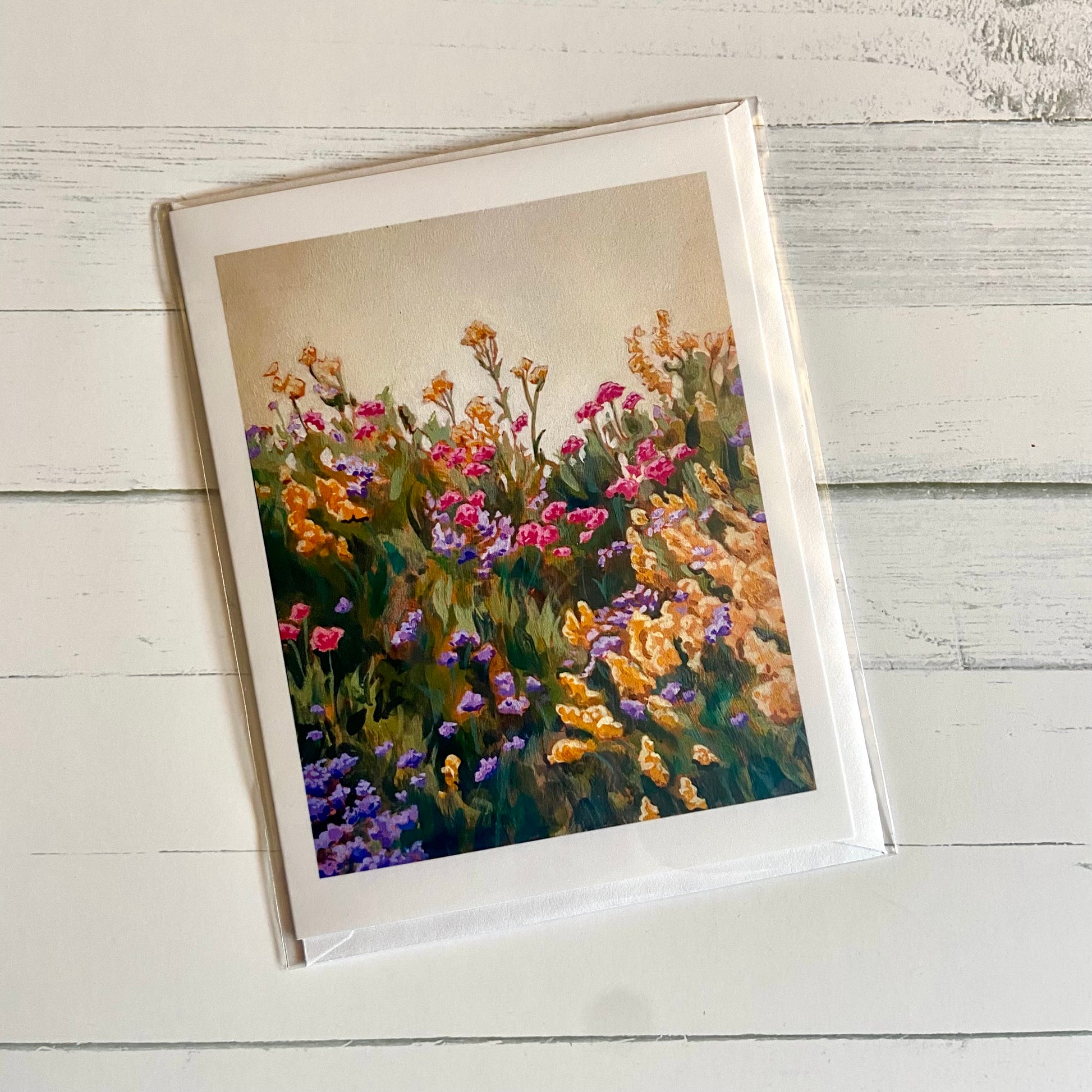 A folded white greeting card printed on the front with lush floral painting, Goldenrod Hill, by artist Jackie Hanson. The top third of the painting is a pale yellow sky and the remainder is a thick, grassy hill with bushy flowers in pink, gold and purple.