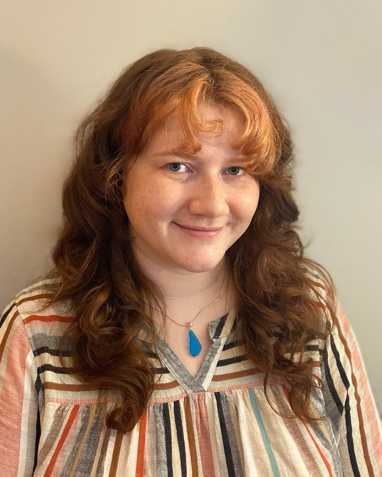 A portrait of a smiling woman looking at the camera. She has red wavy hair and wears a multicolor striped top and a blue necklace.