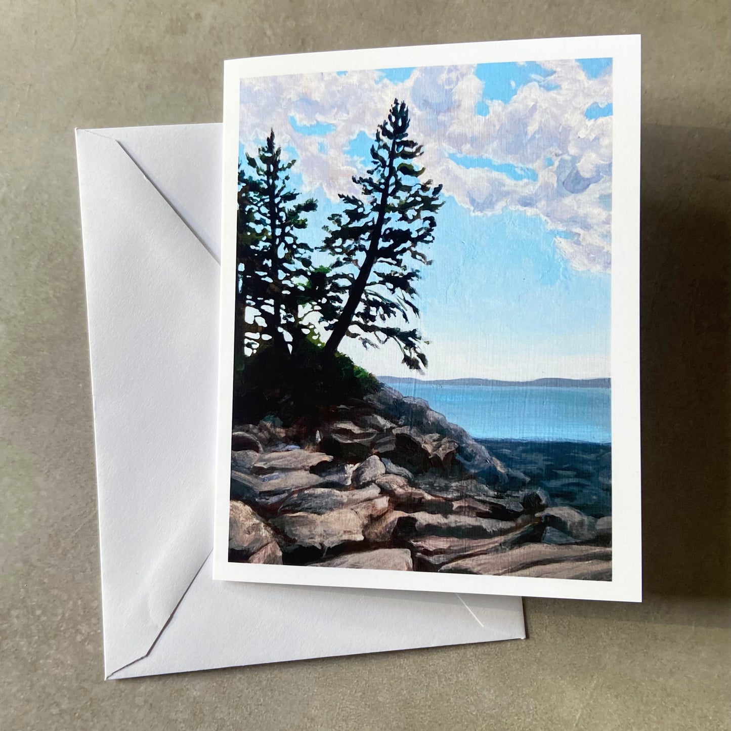 A folded white greeting card and envelope on a grey surface. Depicted on the card is a painting of a clear day by the ocean on a rocky shore, with two pine trees just above the beach.