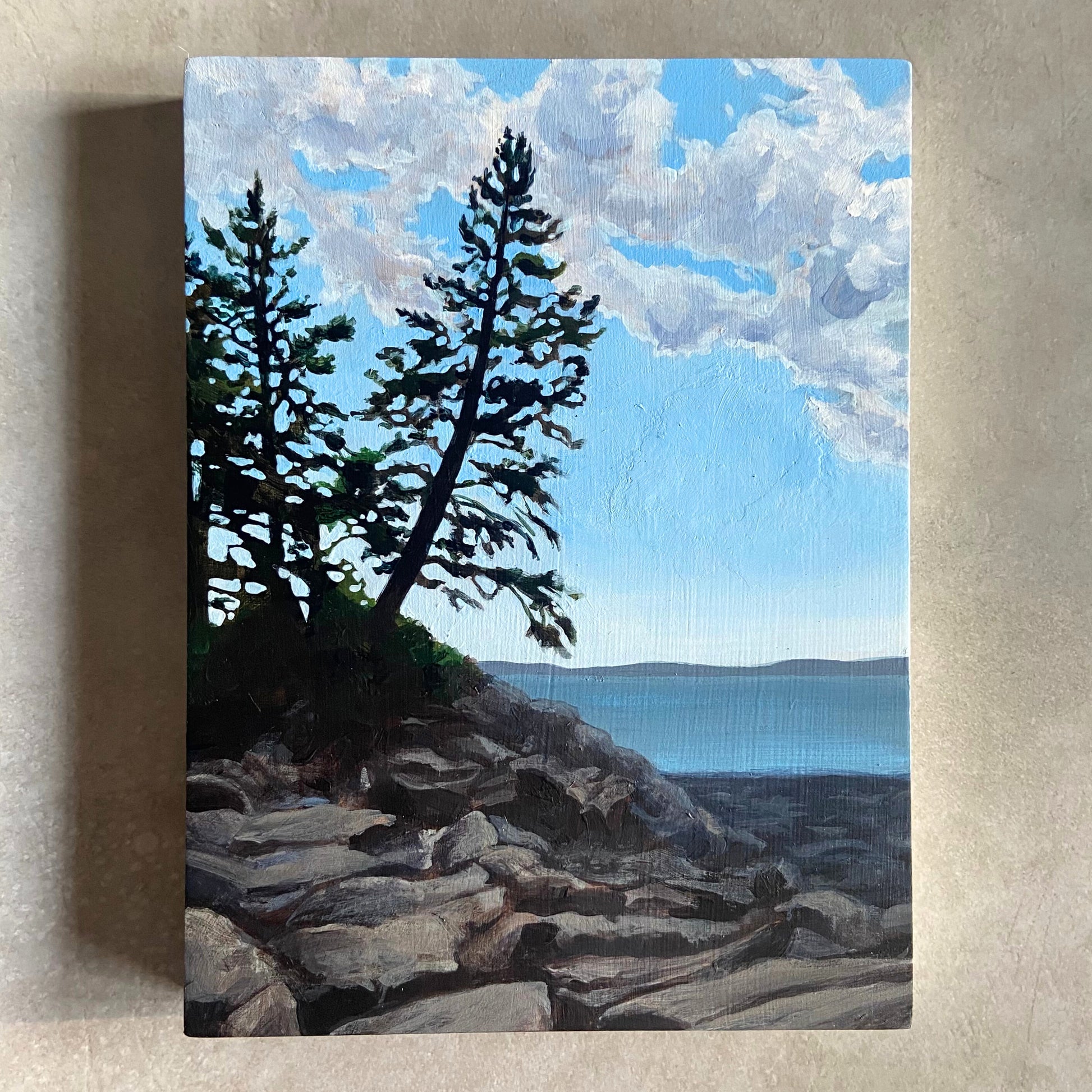 An acrylic landscape painting on wood panel, sitting on a beige, textured background. The artwork, portrait oriented, depicts a couple of pine trees growing above a rocky beach. A calm ocean bay is in the background and the sky is blue with white clouds.