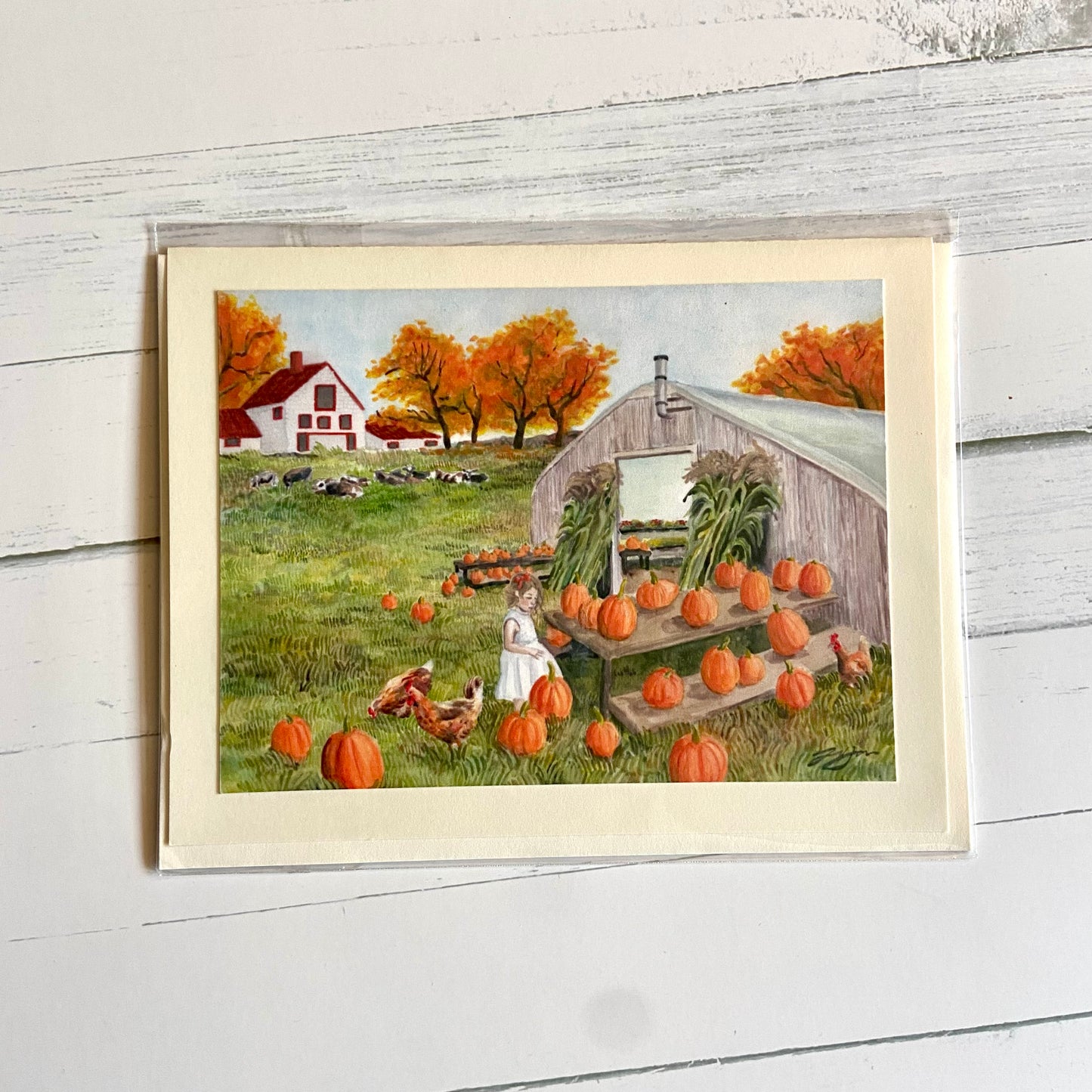 A folded cream-colored greeting card with an image of my painting, Homespun. Shown is a greenhousebuilding with dozens of pumpkins arranged outside. A little girl browses the gourds, accompanied by chickens. Farther away is a white farmhosue with a red roof, autumn orange trees, and a herd of resting cows.