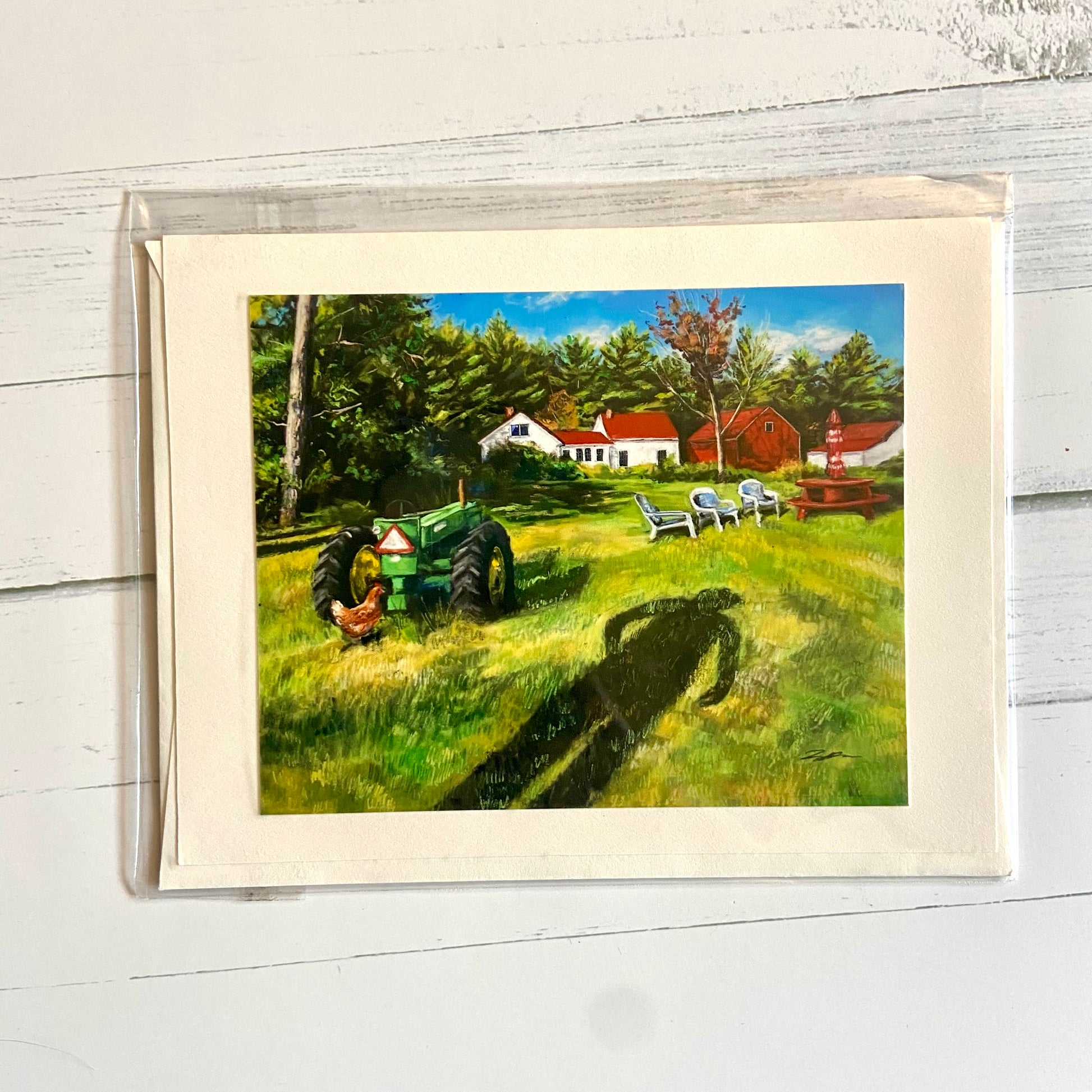 This is a folded cream-colored card depicting my pastel landscape painting, In Between the Field and the Farm. Shown is the shadow of a person extending over a grassy field toward a green tractor, a chicken, and a picnic table with chairs. In the distance is a white farmhouse with a red roof, a red barn, and a thick border of pine trees in back.