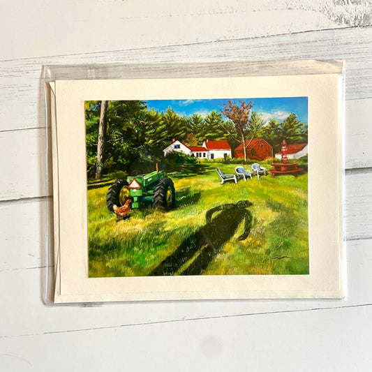 This is a folded cream-colored card depicting my pastel landscape painting, In Between the Field and the Farm. Shown is the shadow of a person extending over a grassy field toward a green tractor, a chicken, and a picnic table with chairs. In the distance is a white farmhouse with a red roof, a red barn, and a thick border of pine trees in back.
