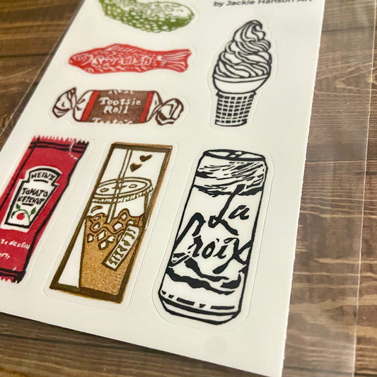A close-up angled view of the eraser art sticker sheet. Individual cut lines around the 7 stickers can be seen. The stickers are each around 1x2 inches and include a pickle, Swedish fish, tootsie roll, ice cream, ketchup, iced coffee, and La Croix.