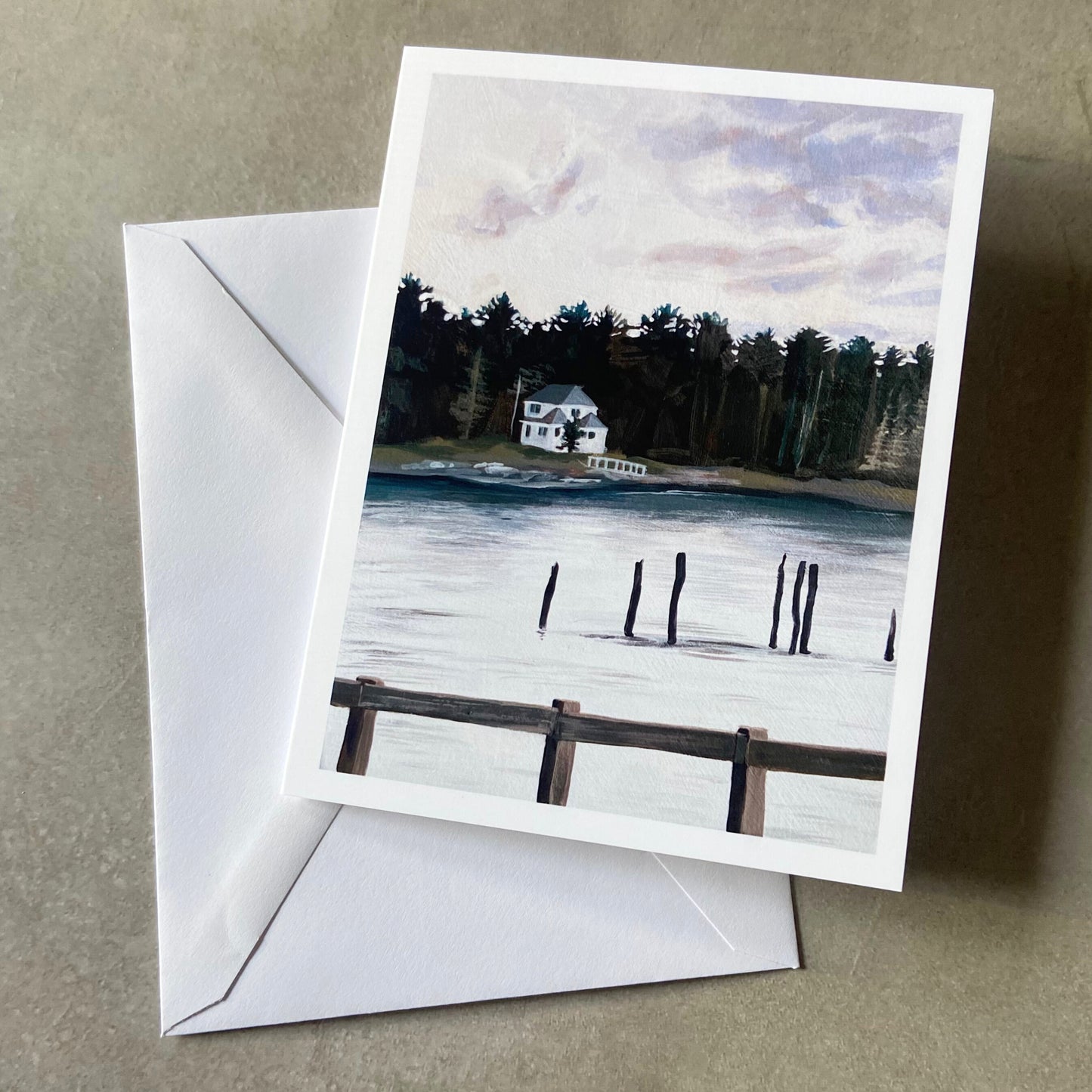 A folded white greeting card and envelope on a grey surface. On the card is a painting of a white house by a body of water on an overcast day. There are the standing remains of a pier in the water and a wooden fence at the bottom.