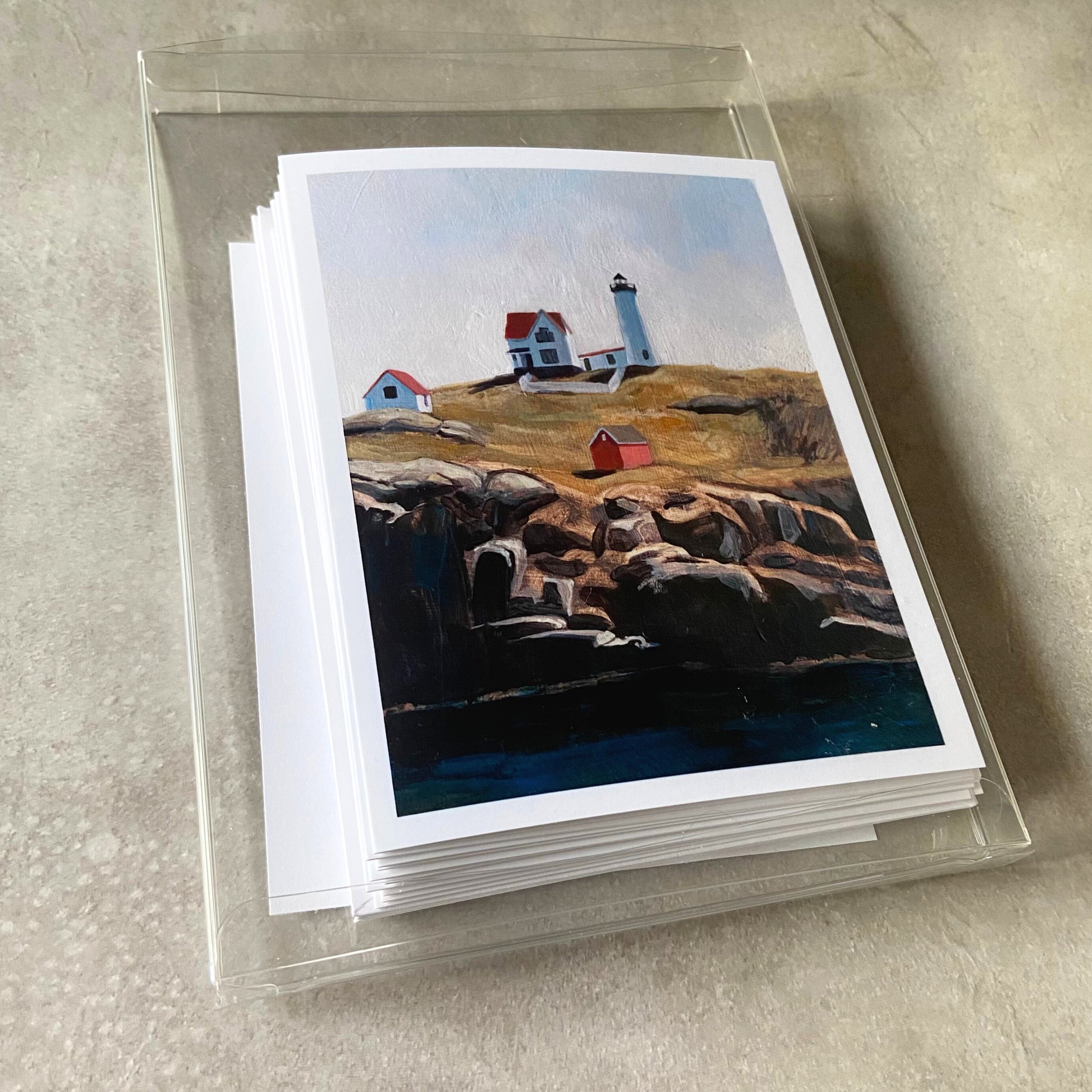 A clear plastic box containing a stack of greeting cards and envelopes. The top card shown depicts a painting of Nubble Lighthouse in York, Maine.
