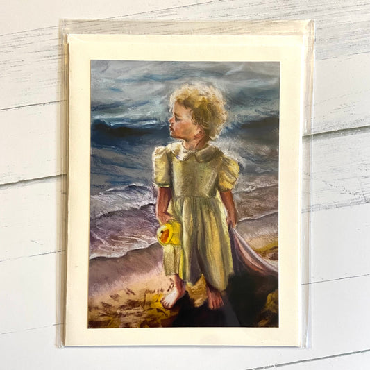 A folded greeting card with a painting of a little girl in a yellow dress standing by the water. She has curly white-blonde hair, holds a stuffed ducky, and is dragging a pink blanket in the sand behind.