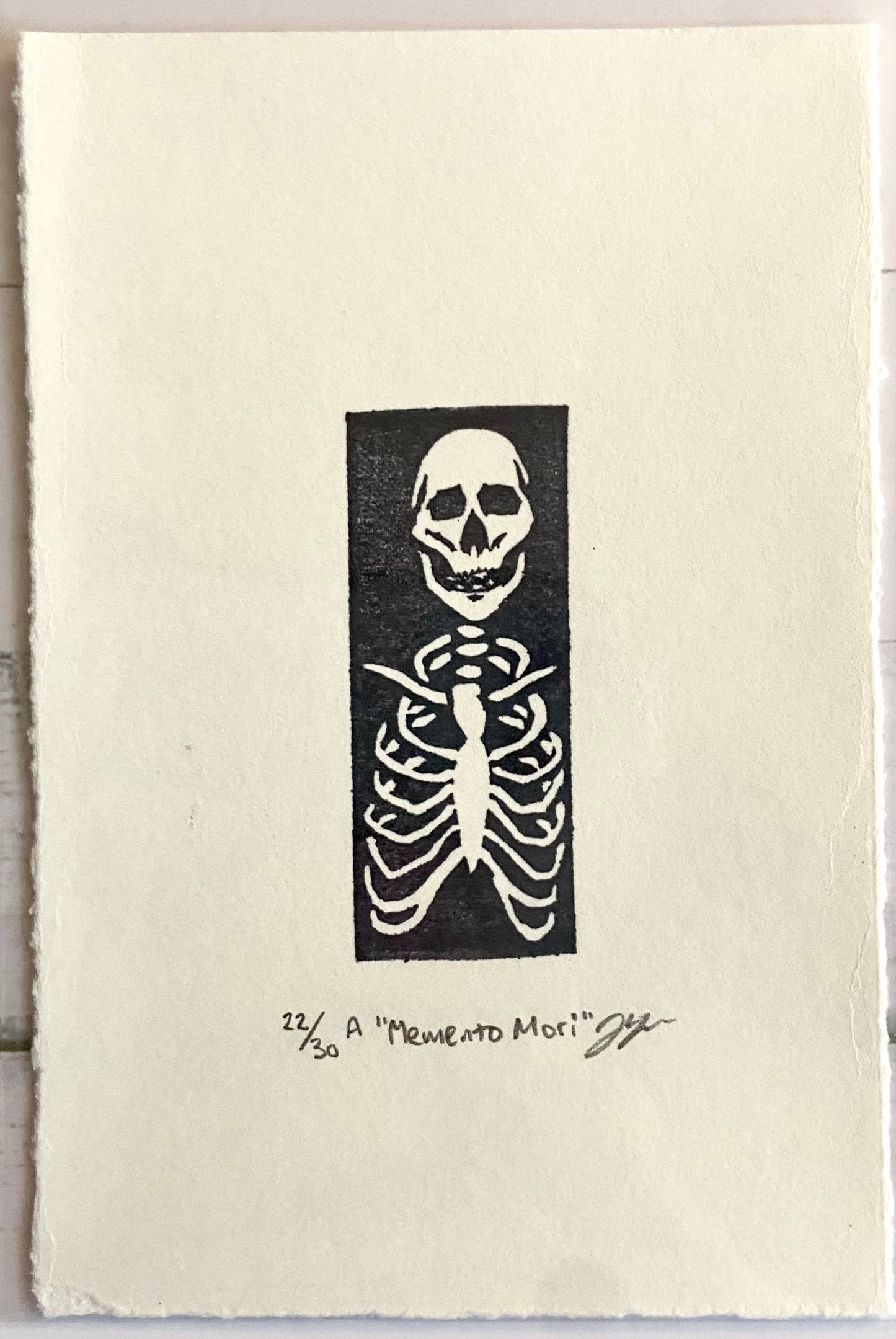 A tiny stamped print showing a skeleton's ribcage and skull in white on a black rectangle, with a wide paper border around it. Eraser printed are made by carving pink erasers and stamping them. The print is titled "Memento Mori" and is signed and editioned.