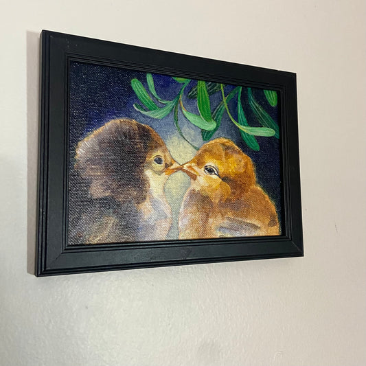 A small painting of two baby chicks beak-to-beak under mistletoe leaves with a deep blue background. The art is framed in black and hangs on a light gray wall.