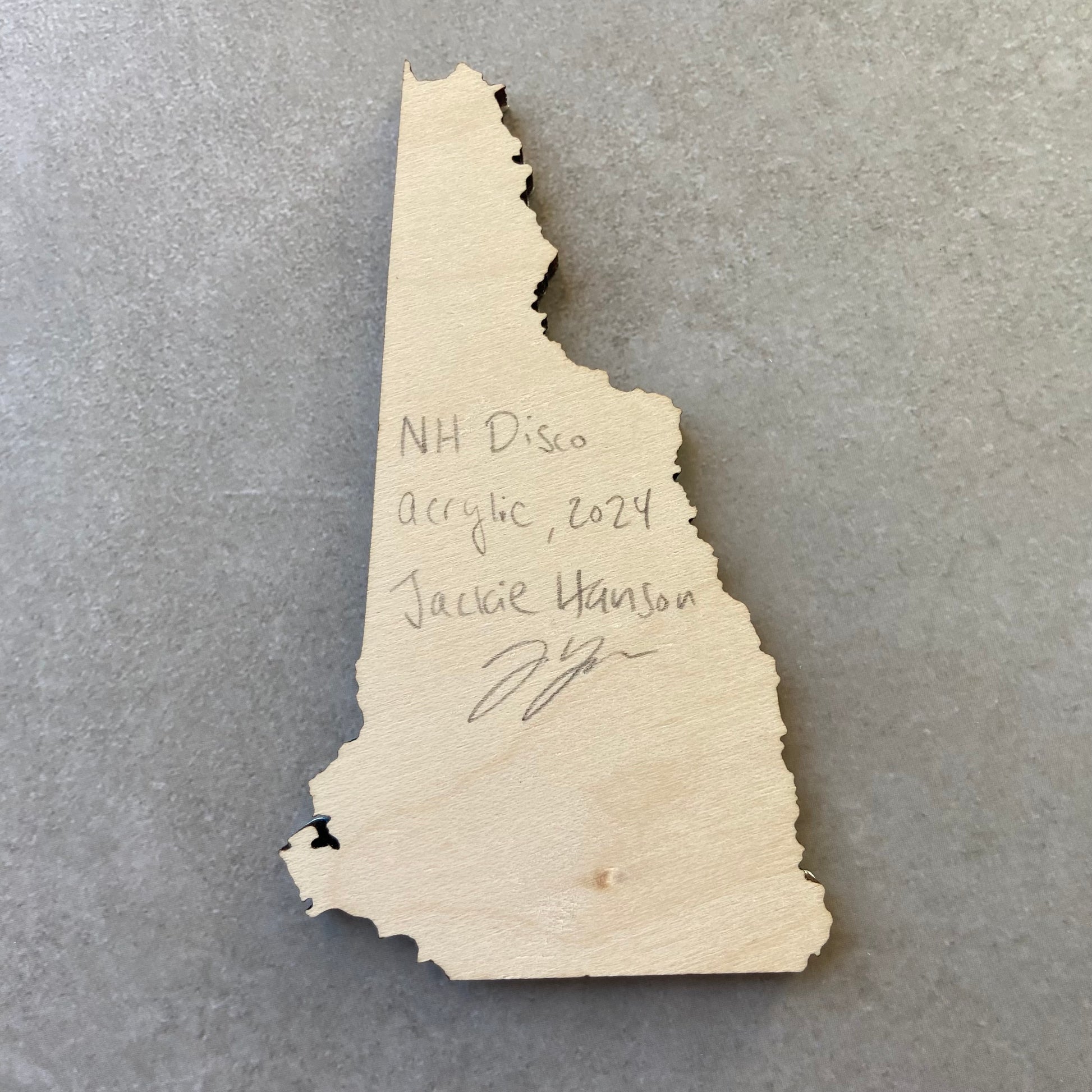 The back of a New Hampshire-shaped painting on wood. Writing on the bare wood in pencil reads, "NH Disco. Acrylic, 2024. Jackie Hanson" and signed by the artist.