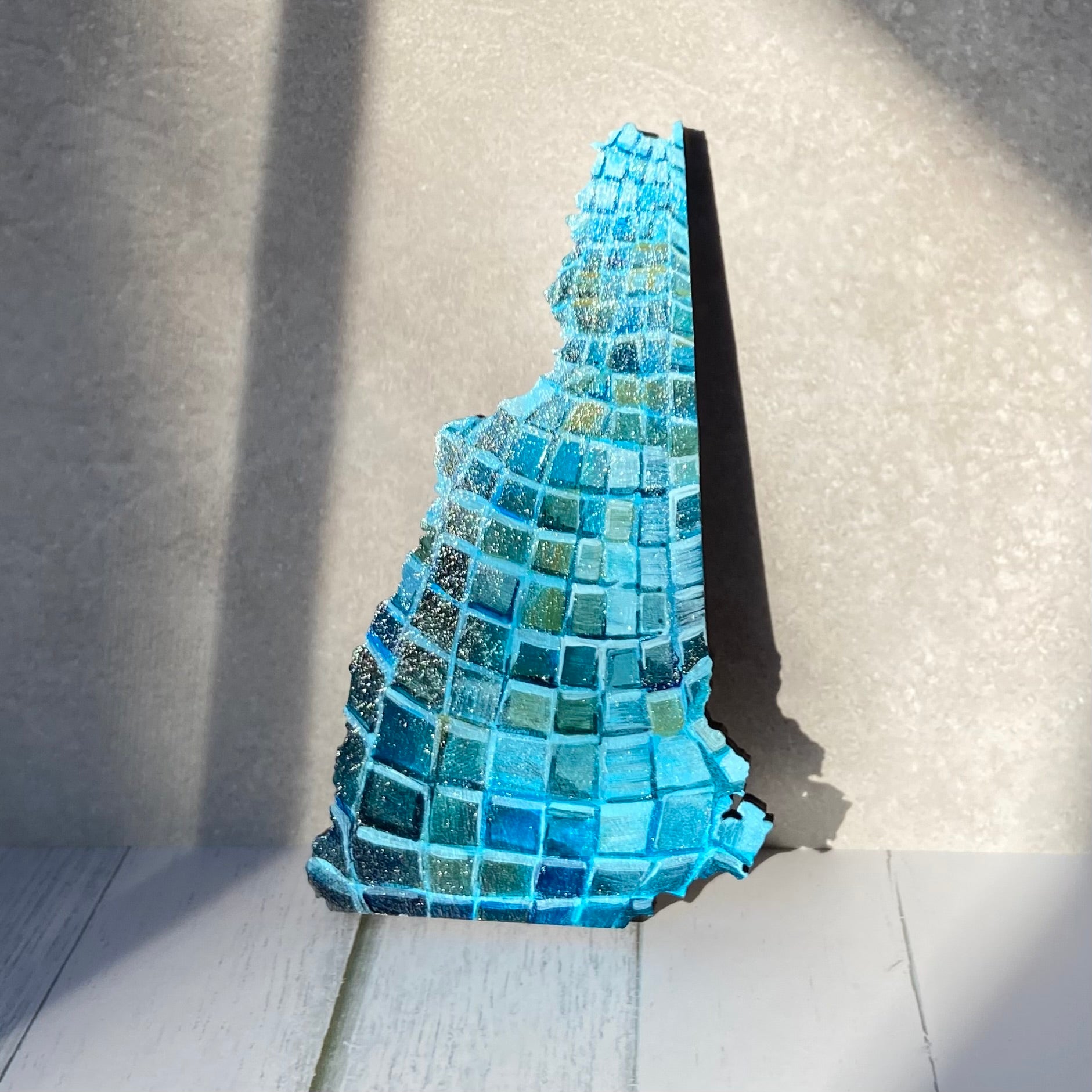 A New Hampshire- shaped painting on wood, painted with the pattern of a blue disco ball. The painting is stood up in a patch of sunlight.