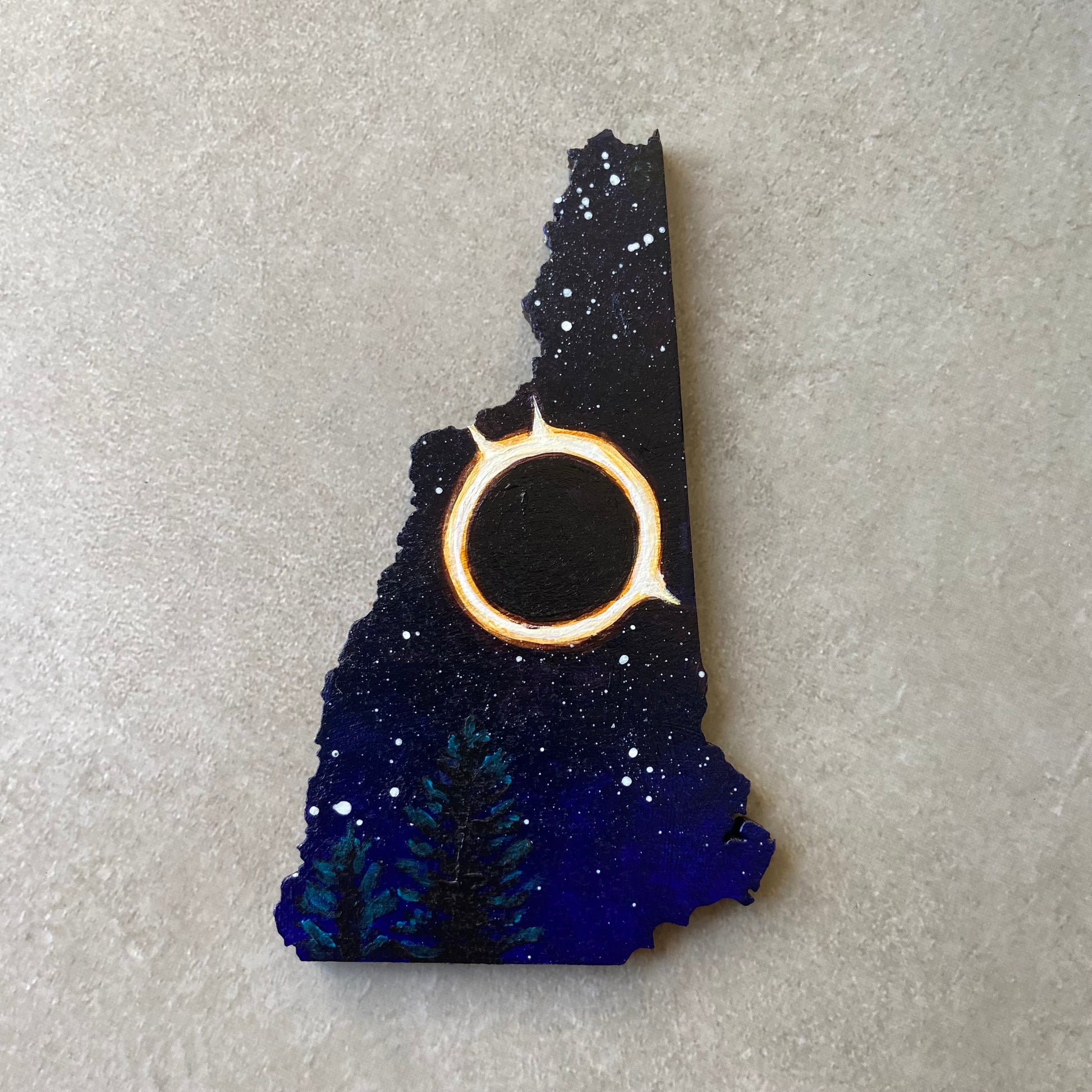 A New Hampshire-shaped painting of a starry night sky with a fiery bright ring in the center. Below are some pine trees.