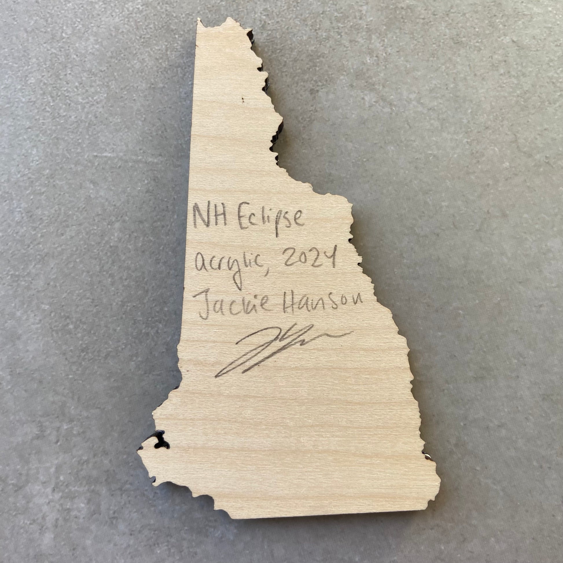 The back of a New Hampshire-shaped painting on wood. Writing on the bare wood in pencil reads, "NH Eclipse. Acrylic, 2024. Jackie Hanson" and signed by the artist.