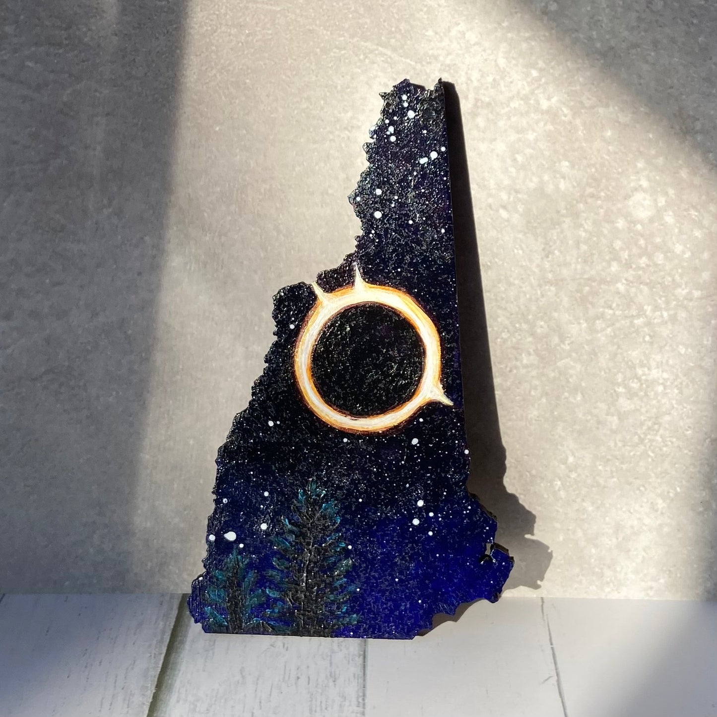 A New Hampshire-shaped painting of a starry night sky with a fiery bright ring in the center. Below are some pine trees. The painting is standing upright in a patch of sunlight.