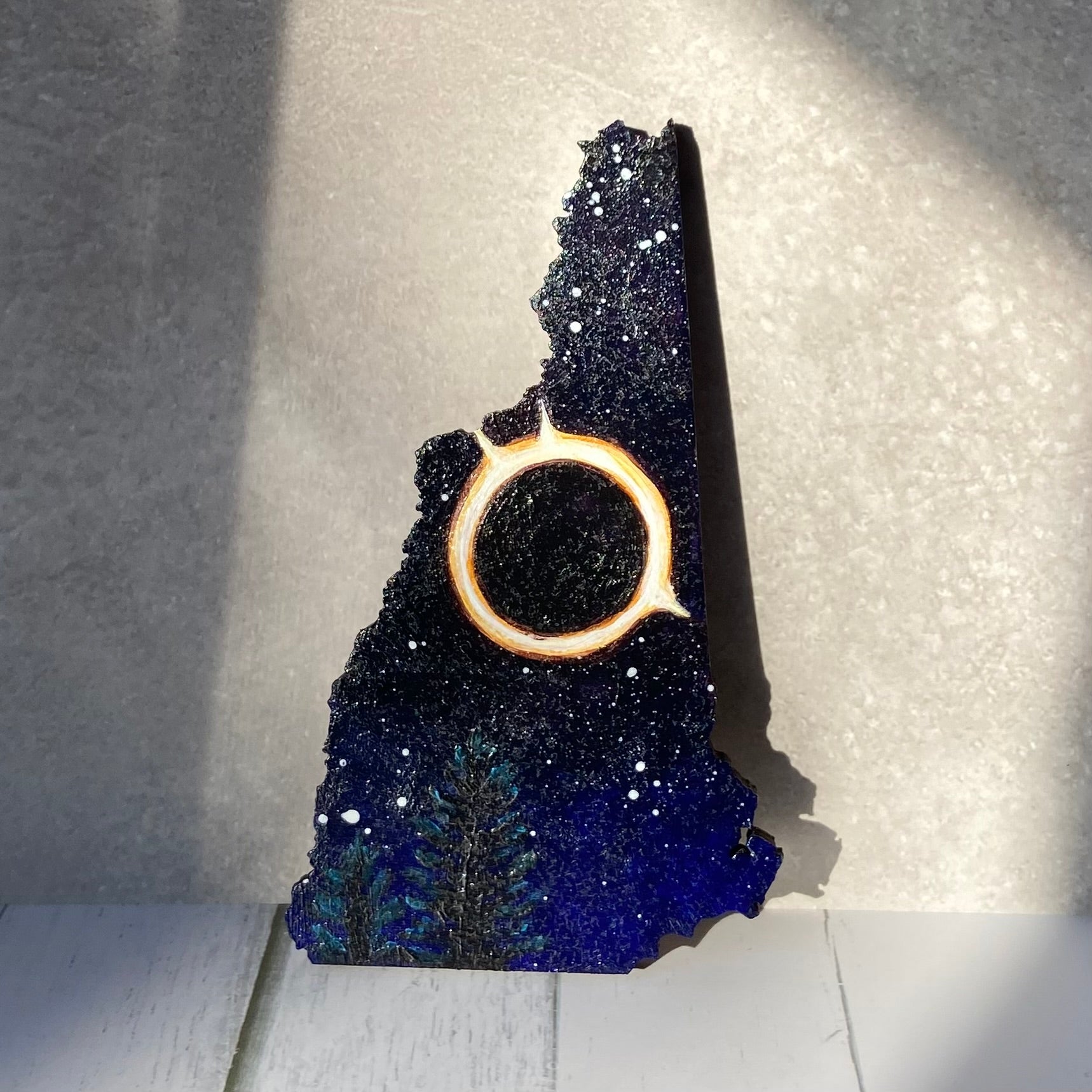 A New Hampshire-shaped painting of a starry night sky with a fiery bright ring in the center. Below are some pine trees. The painting is standing upright in a patch of sunlight.