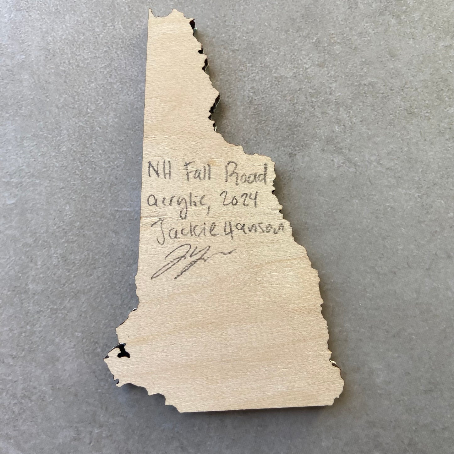 The back of a New Hampshire-shaped painting on wood. Writing on the bare wood in pencil reads, "NH Fall Road. Acrylic, 2024. Jackie Hanson" and signed by the artist.
