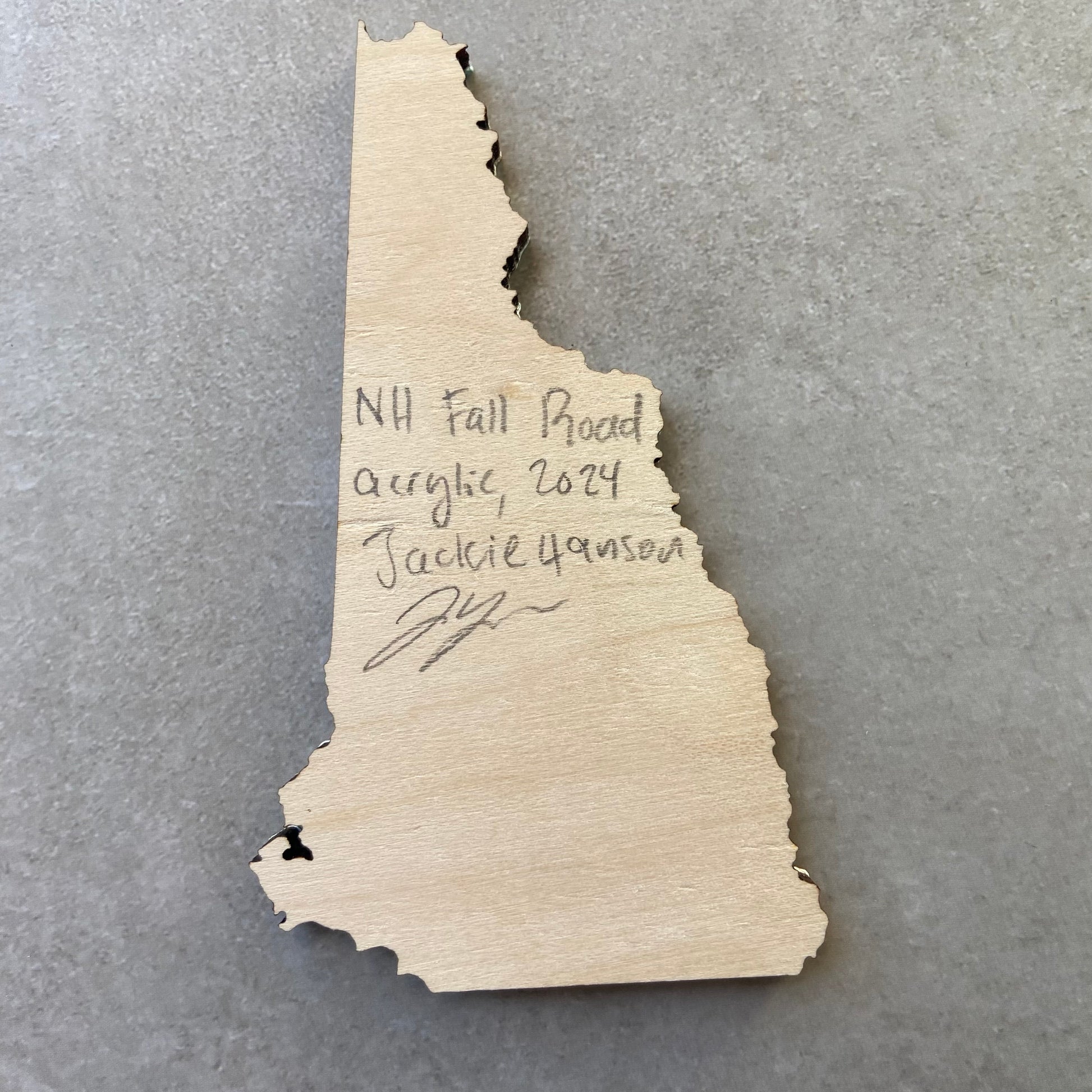 The back of a New Hampshire-shaped painting on wood. Writing on the bare wood in pencil reads, "NH Fall Road. Acrylic, 2024. Jackie Hanson" and signed by the artist.