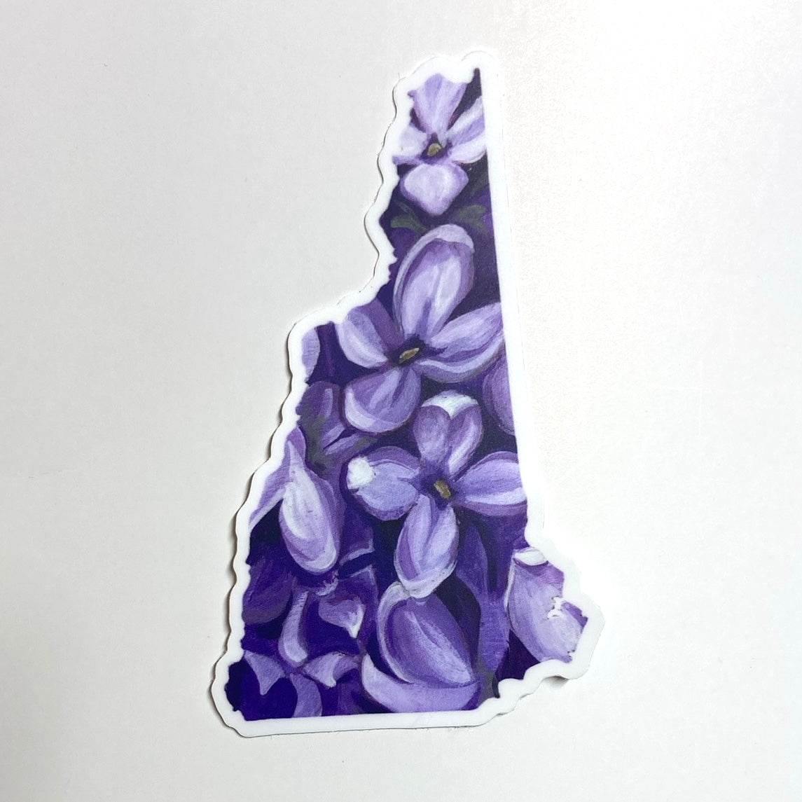 A sticker in the shape of New Hampshire with a painting of purple lilac flowers.