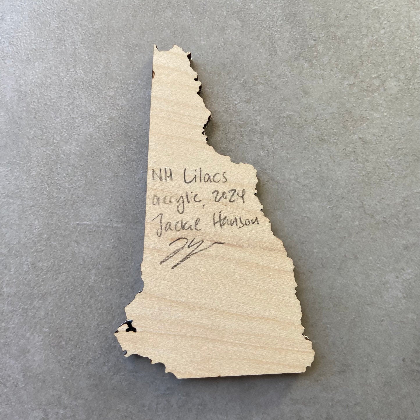 The back of a New Hampshire-shaped painting on wood. Writing on the bare wood in pencil reads, "NH Lilacs. Acrylic, 2024. Jackie Hanson" and signed by the artist.