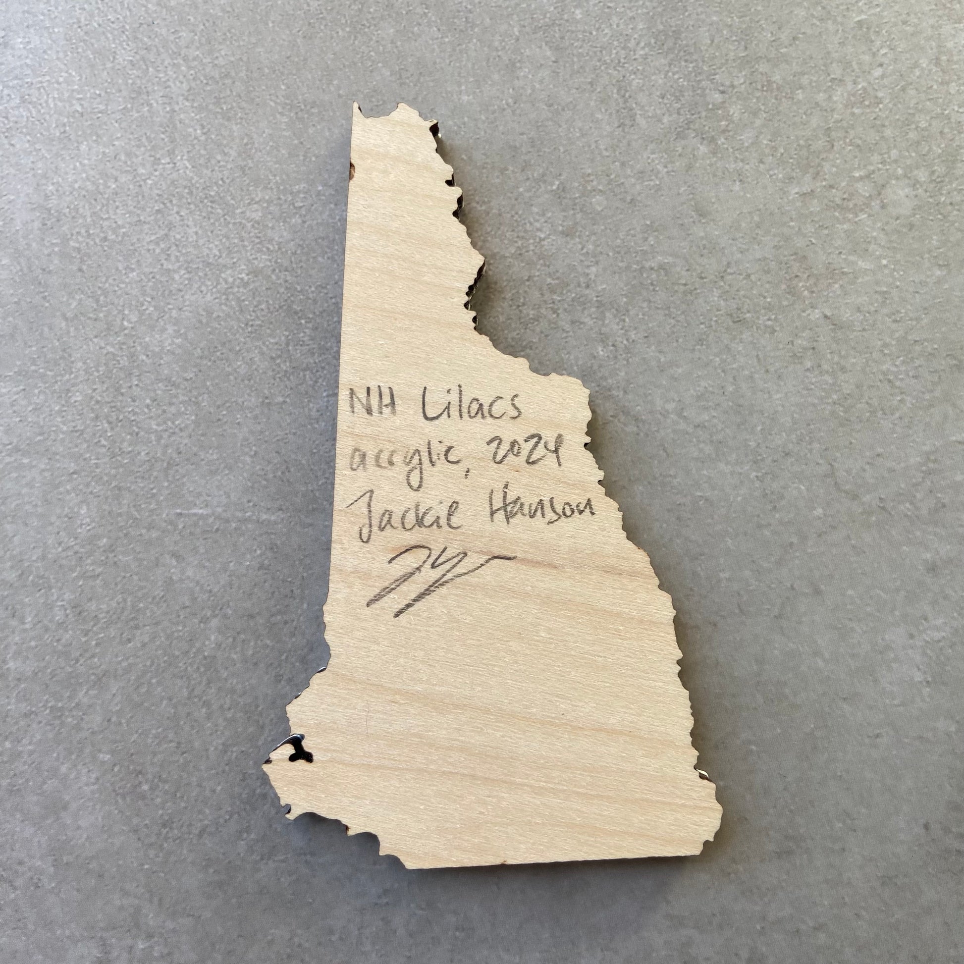 The back of a New Hampshire-shaped painting on wood. Writing on the bare wood in pencil reads, "NH Lilacs. Acrylic, 2024. Jackie Hanson" and signed by the artist.