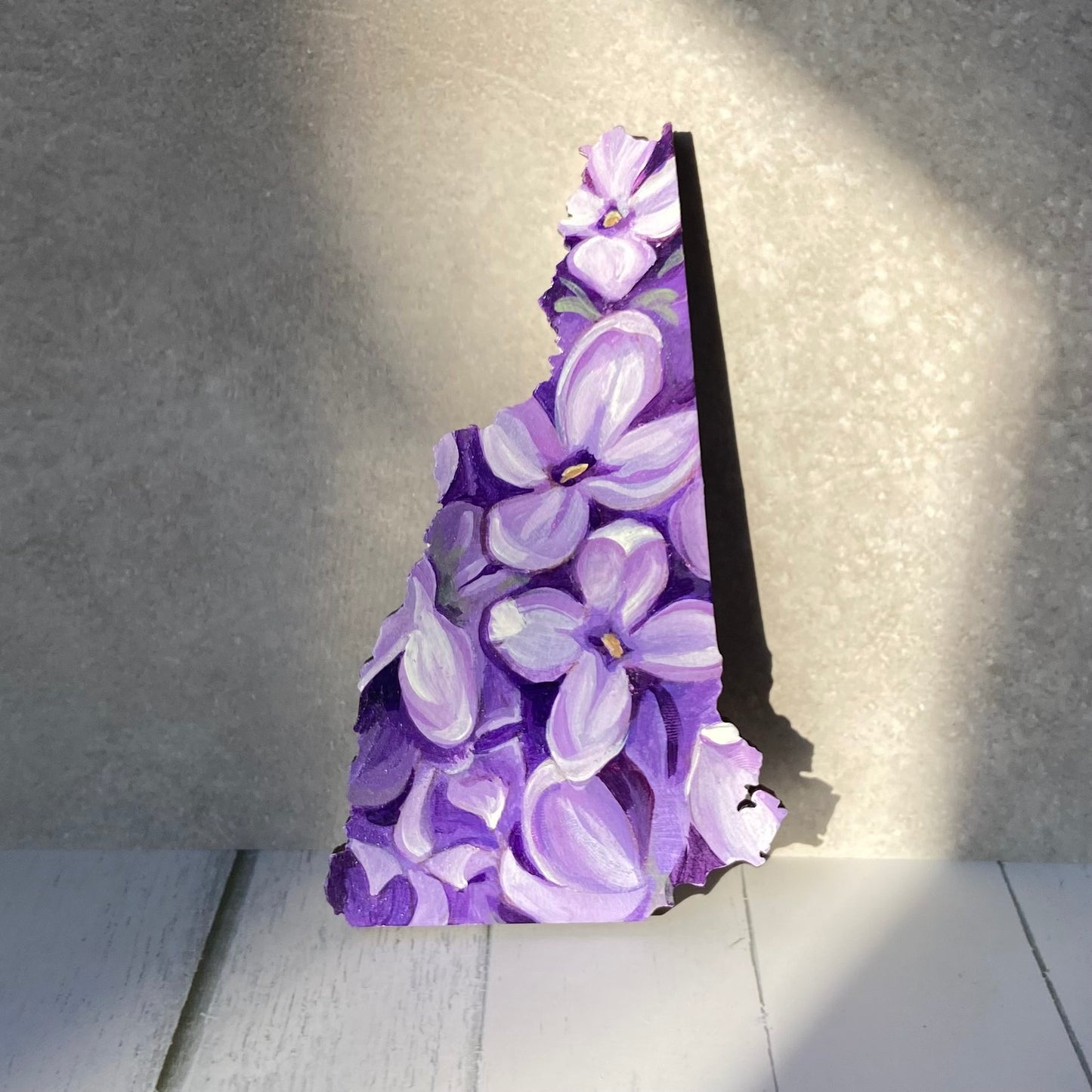 A New Hampshire-shaped painting depicting a close-up look at several blooming purple lilacs. The painting is propped upright in a sunny spot.