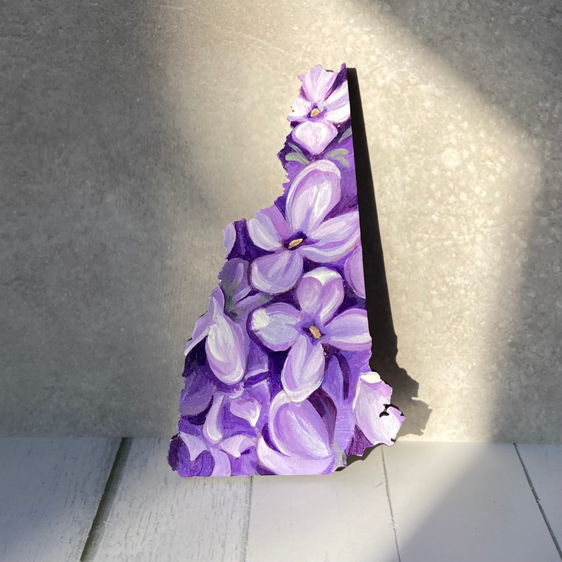 A New Hampshire-shaped painting depicting a close-up look at several blooming purple lilacs. The painting is propped upright in a sunny spot.