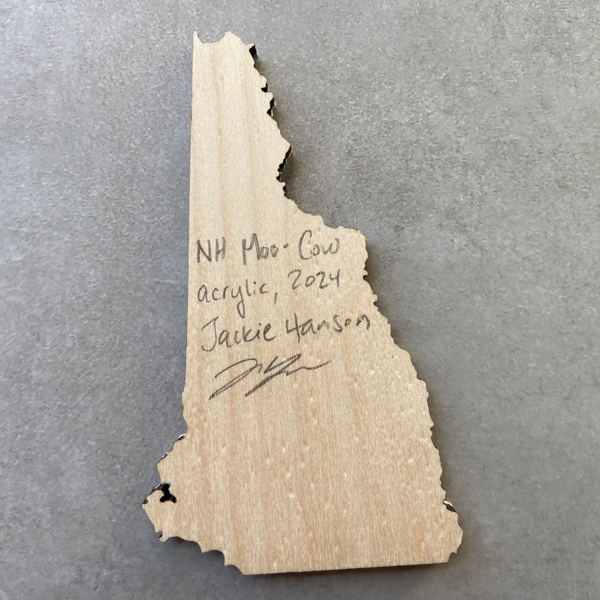 The back of a New Hampshire-shaped painting on wood. Writing on the bare wood in pencil reads, "NH Moo-Cow. Acrylic, 2024. Jackie Hanson" and signed by the artist.