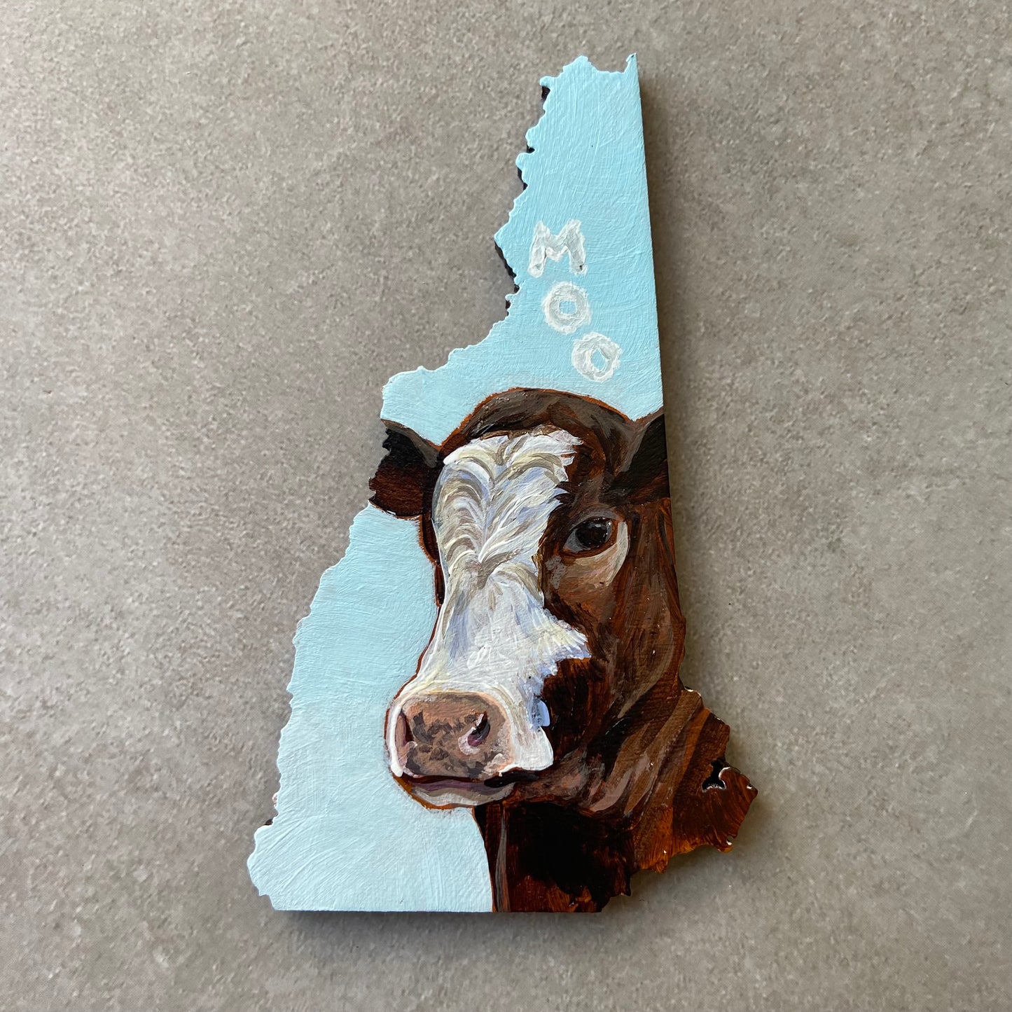 A New Hampshire-shaped painting depicting a brown and white cow's face looking at the viewer. The sky-blue background has three letter-shaped clouds over the cow's head, reading "MOO".